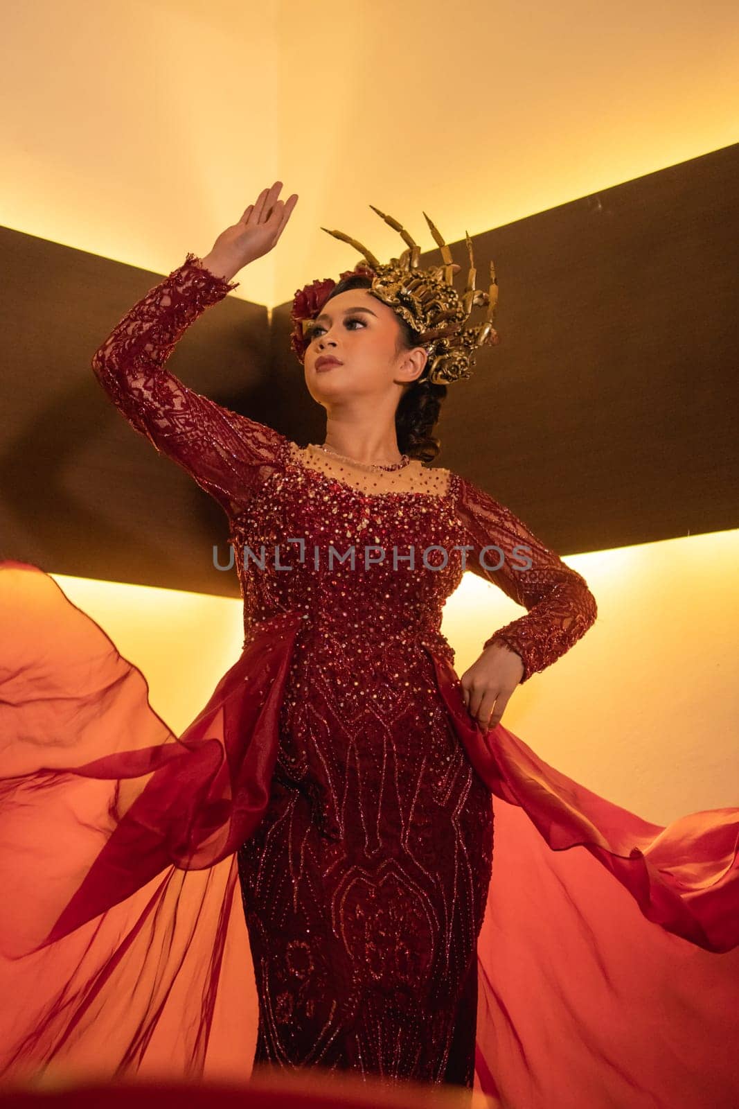 A glamor woman poses with a flying red dress on her body while wearing a gold crown inside the cream studio
