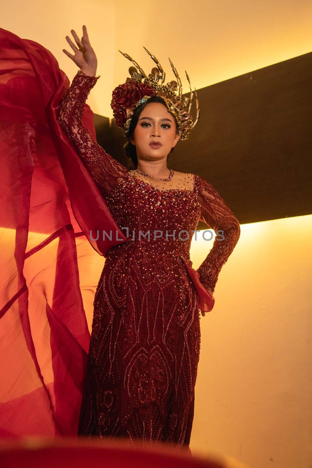 A glamor woman poses with a flying red dress on her body while wearing a gold crown inside the cream studio