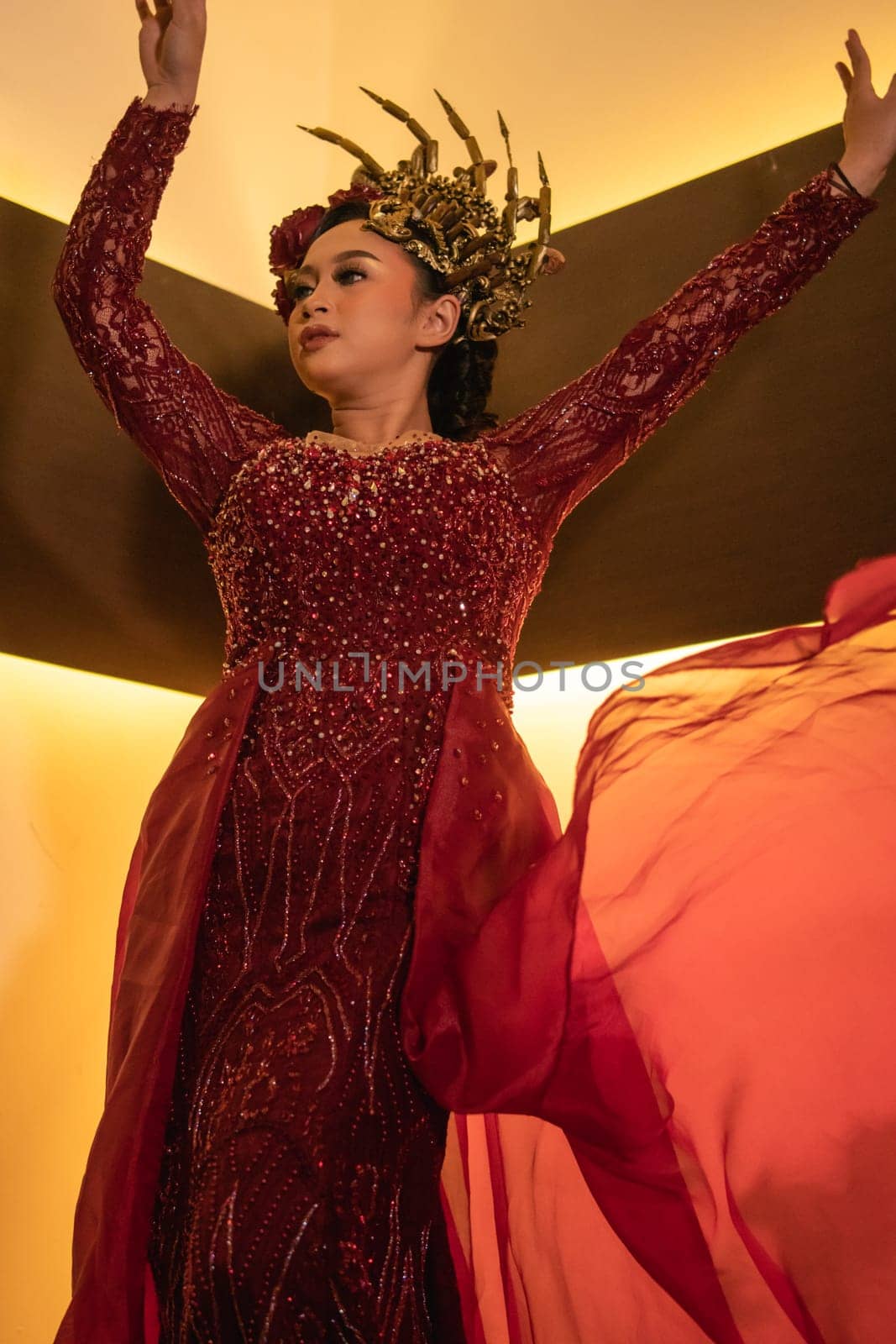 A glamor woman poses with a flying red dress on her body while wearing a gold crown inside the cream studio