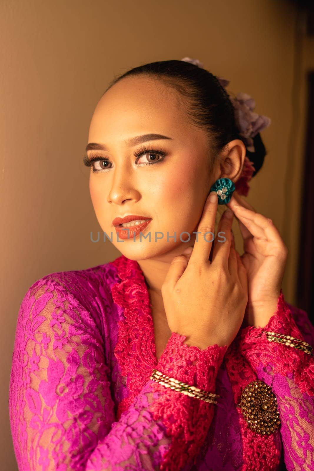Asian woman in a pink dress trying to wear a green earring in her ears inside the studio