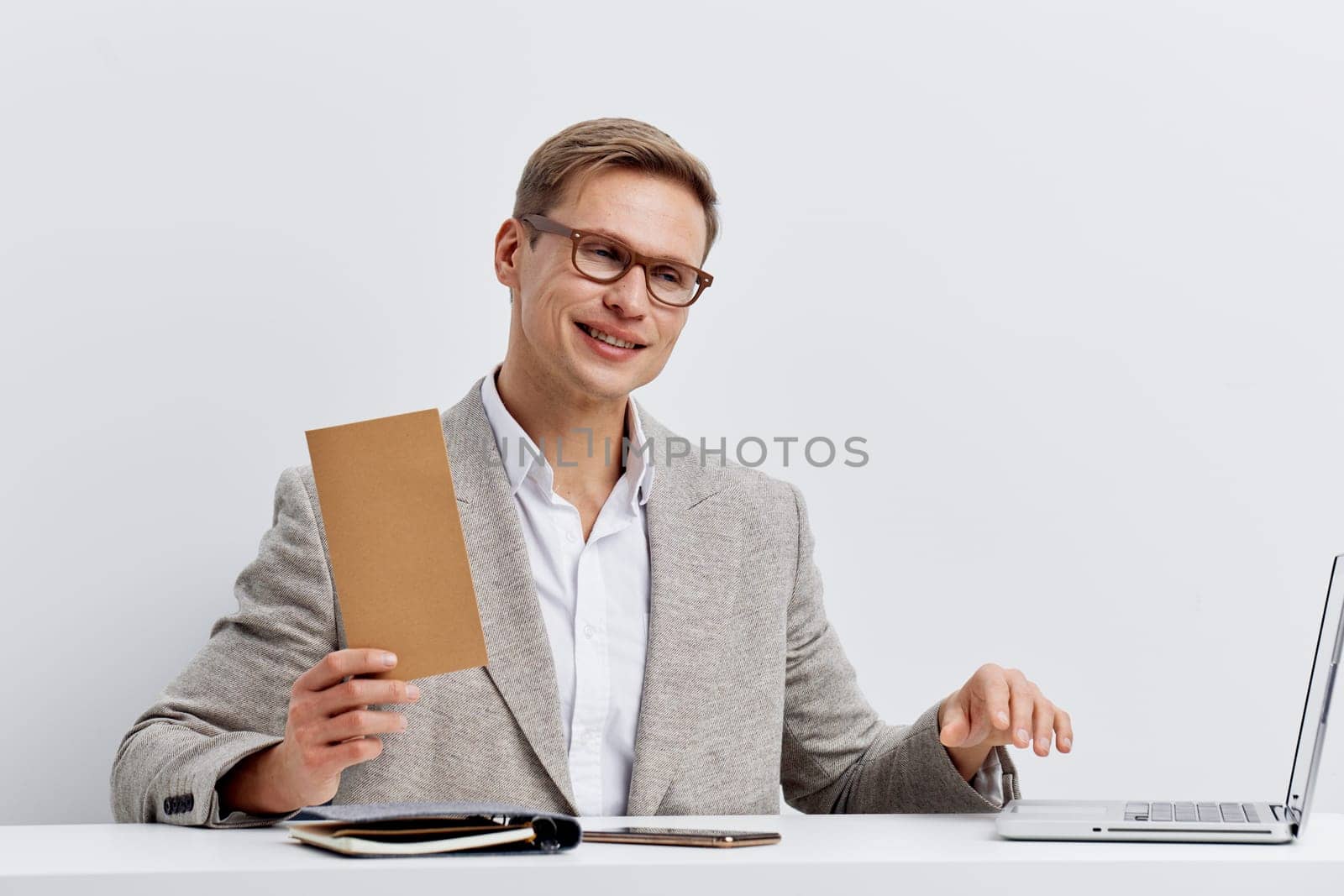 Confident smiling man wearing glasses, grey blazer, and white shirt, working at his desk with a laptop and documents on a minimalist white background. Concept: professional success and modern work environment, office routine