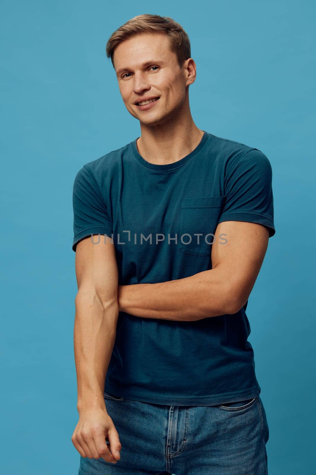 Happy young man with short hair in casual teal t-shirt posing with crossed arms and smiling at the camera on a solid blue background, studio setting, lifestyle image capturing positivity and confidence