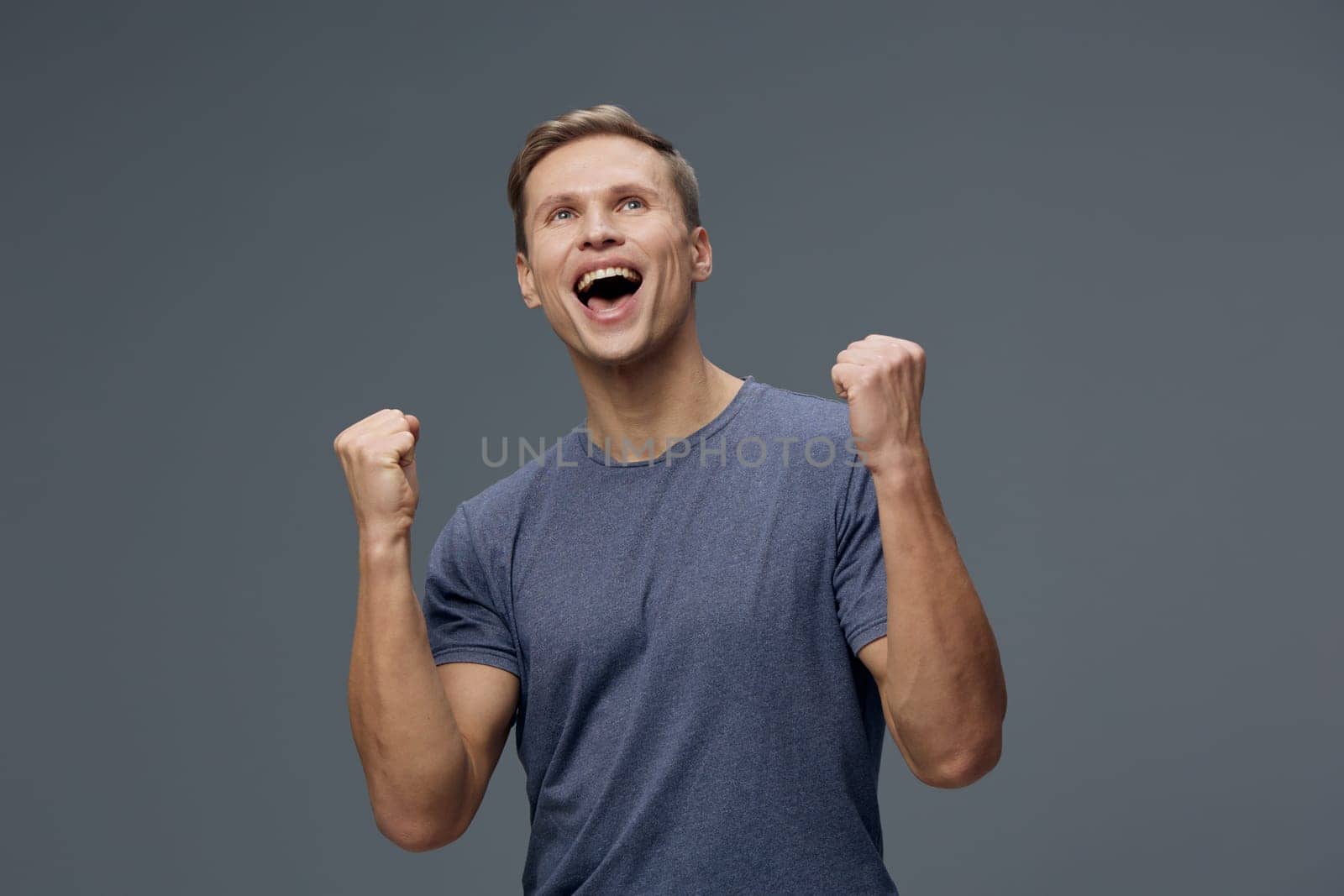 Happy man feeling victorious and energized, casual outfit, smiling with enthusiasm, celebrating achievement, plain gray backdrop, studio environment, positive expression. Concept: success and happiness.