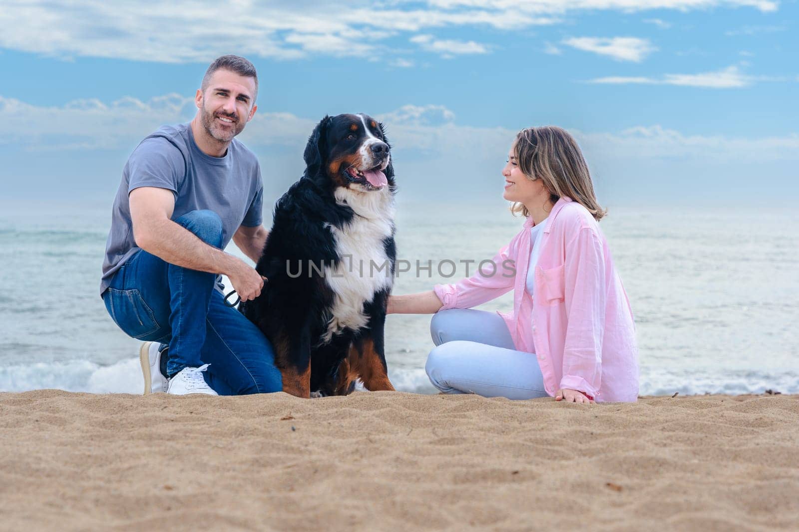 Portrait of a smiling caucasian couple with their large dog on the beach during vacation. Relaxed and joyful moment by the sea, with all three looking at the camera on a bright day.