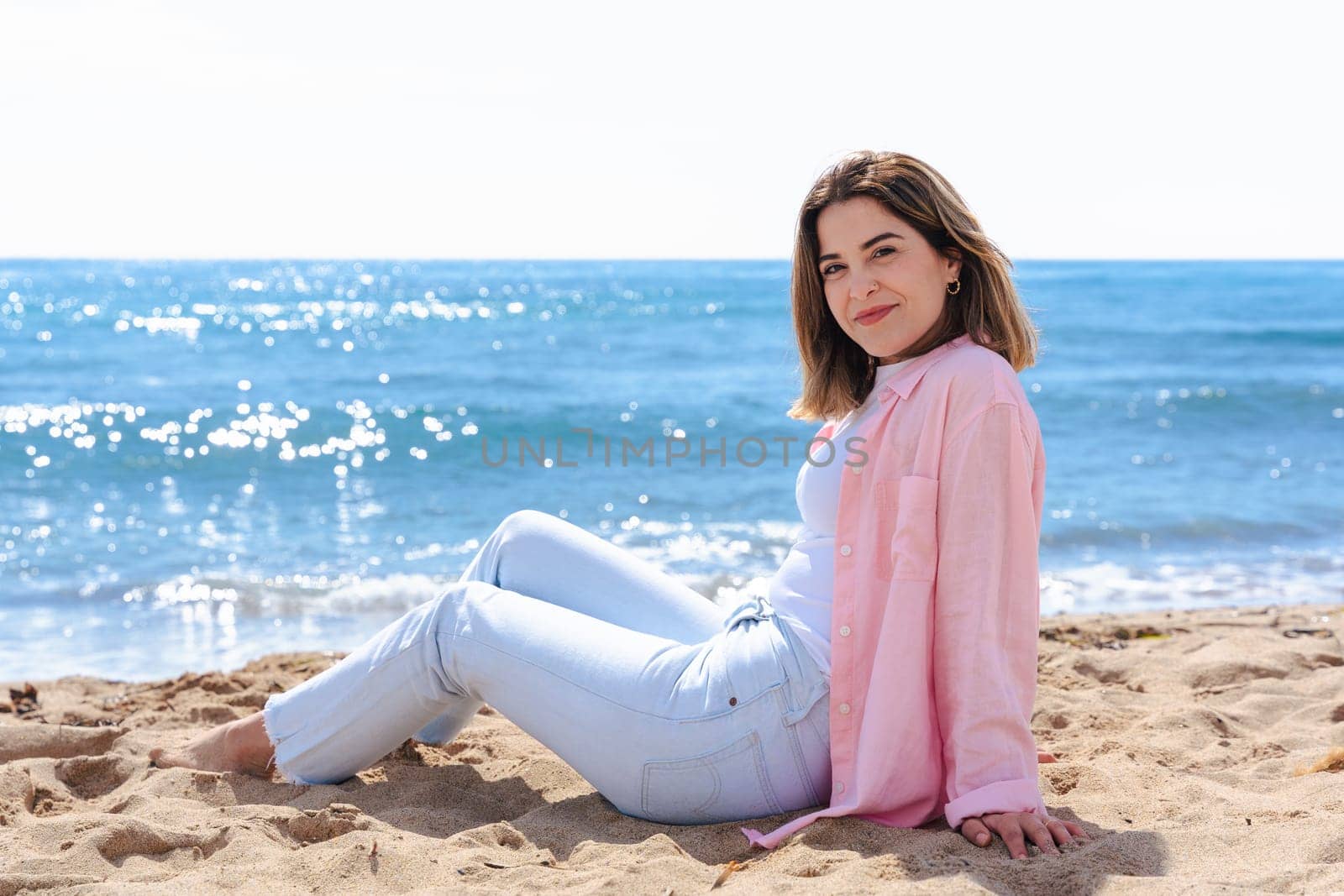 Young caucasian woman on vacation relaxing by the sea, sitting on the beach with a smile. Dressed in casual clothes, enjoying the sunny weather, ocean breeze, and peaceful summer atmosphere.