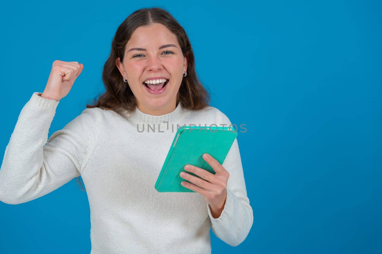 Smiling young woman in white sweater holding a green tablet and raising her fist in celebration while looking at the camera, standing against blue background with copy space, symbolizing success and emotion.