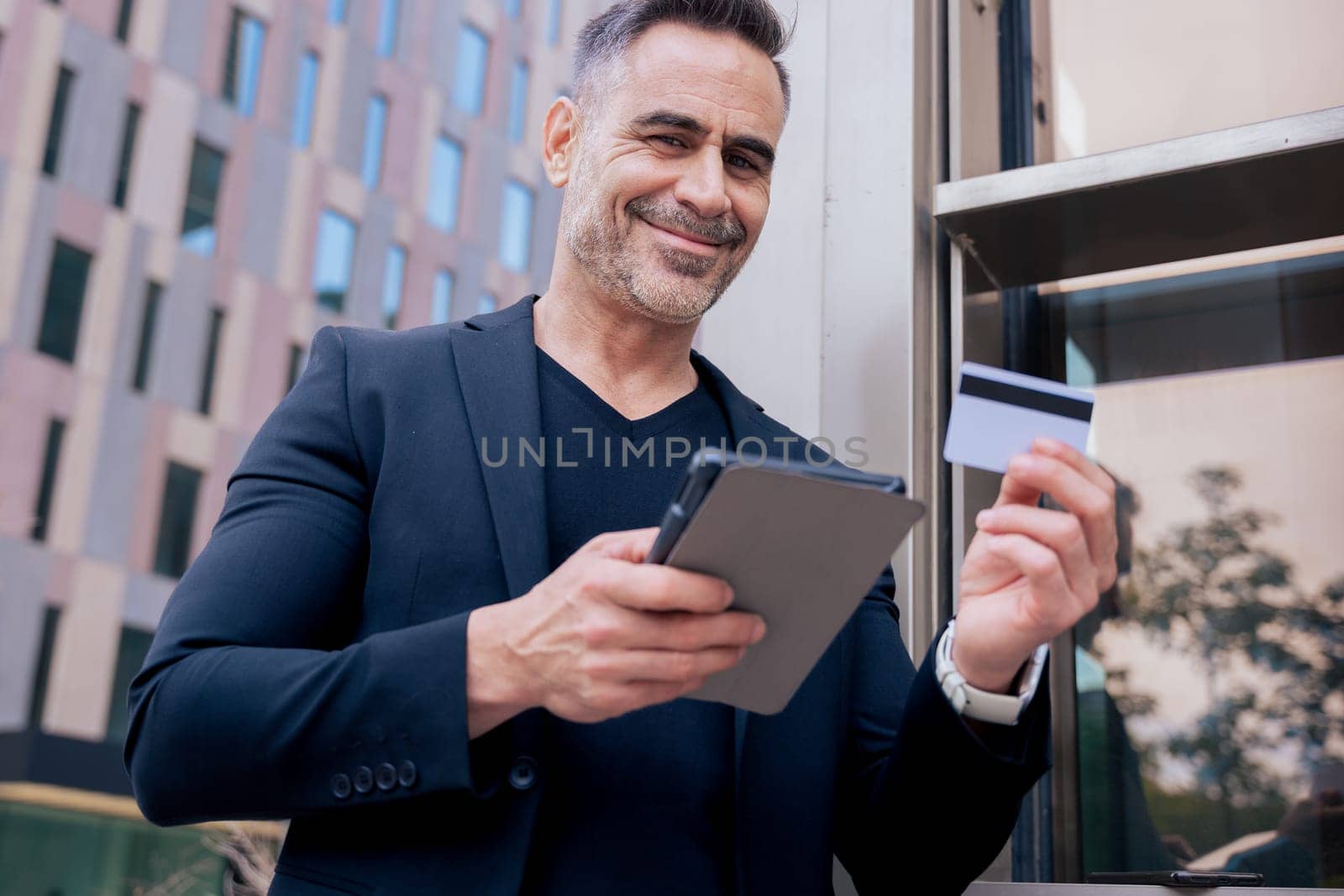 Confident caucasian man with headphones smiling while using tablet and holding a credit card outdoors. Concept of online shopping, digital payment, modern lifestyle, and financial technology.