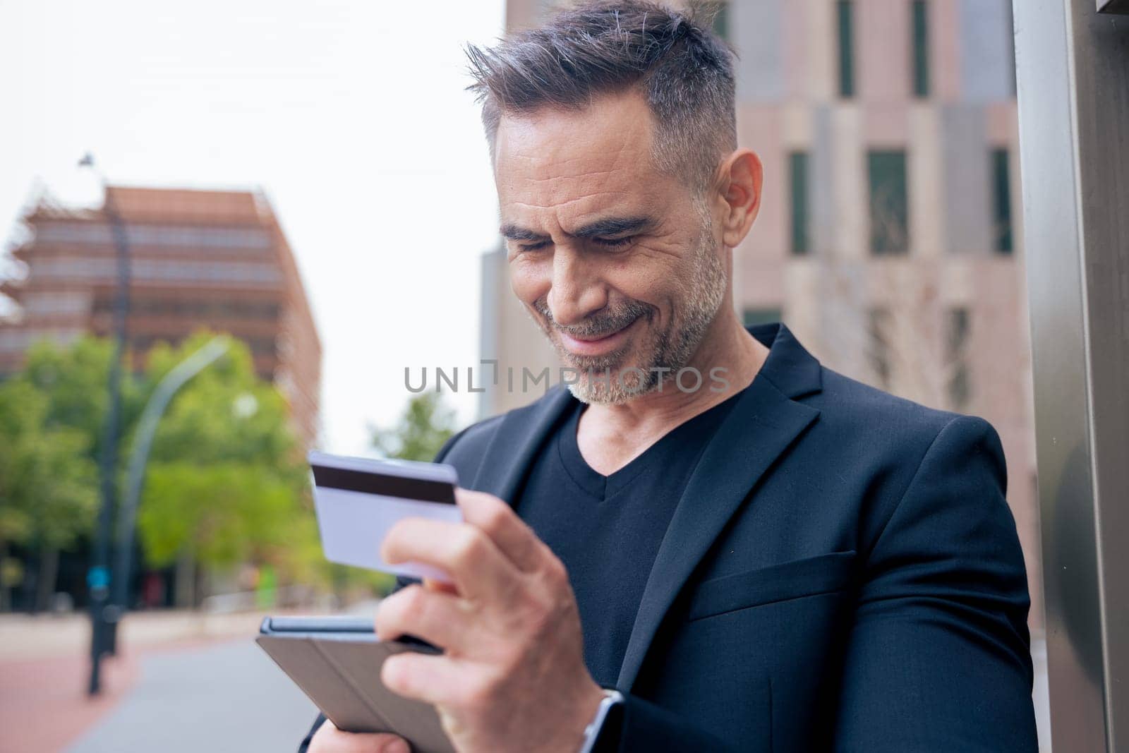 Confident caucasian man with headphones smiling while using tablet and holding a credit card outdoors. Concept of online shopping, digital payment, modern lifestyle, and financial technology.