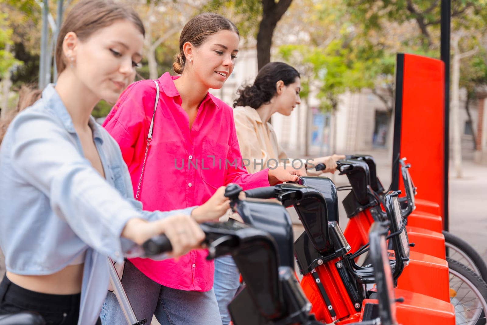 Happy young women renting bicycles in an urban park, smiling and preparing for a fun ride. Dressed casually, they enjoy a lively outdoor setting surrounded by trees and modern city buildings.