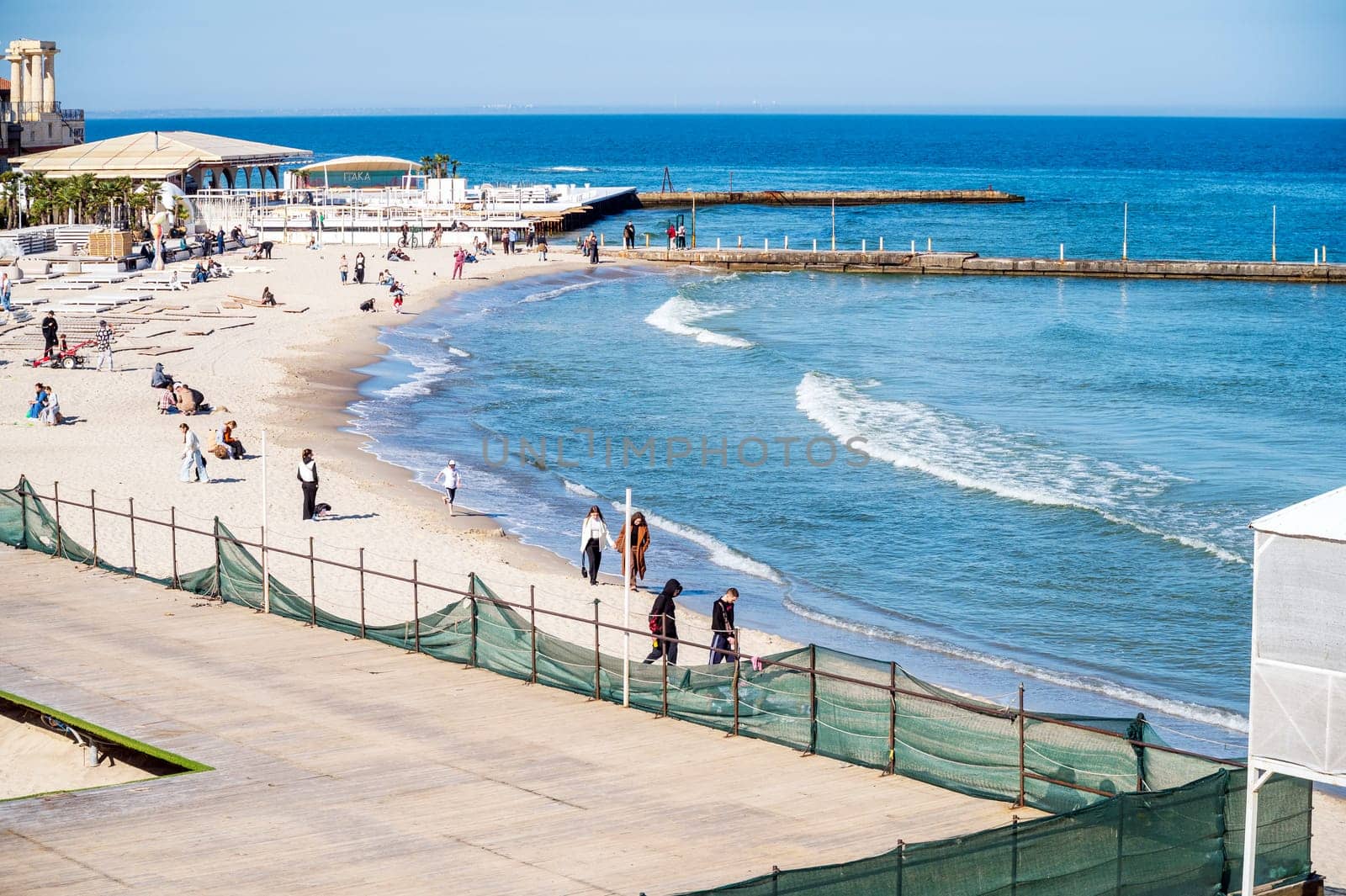 Odessa, Ukraine - April 19, 2025: City beach in Arcadia. Sea sand beach, people sitting on the beach and walking. Sandy beach in the midday sun. Concrete piers go into the calm sea.