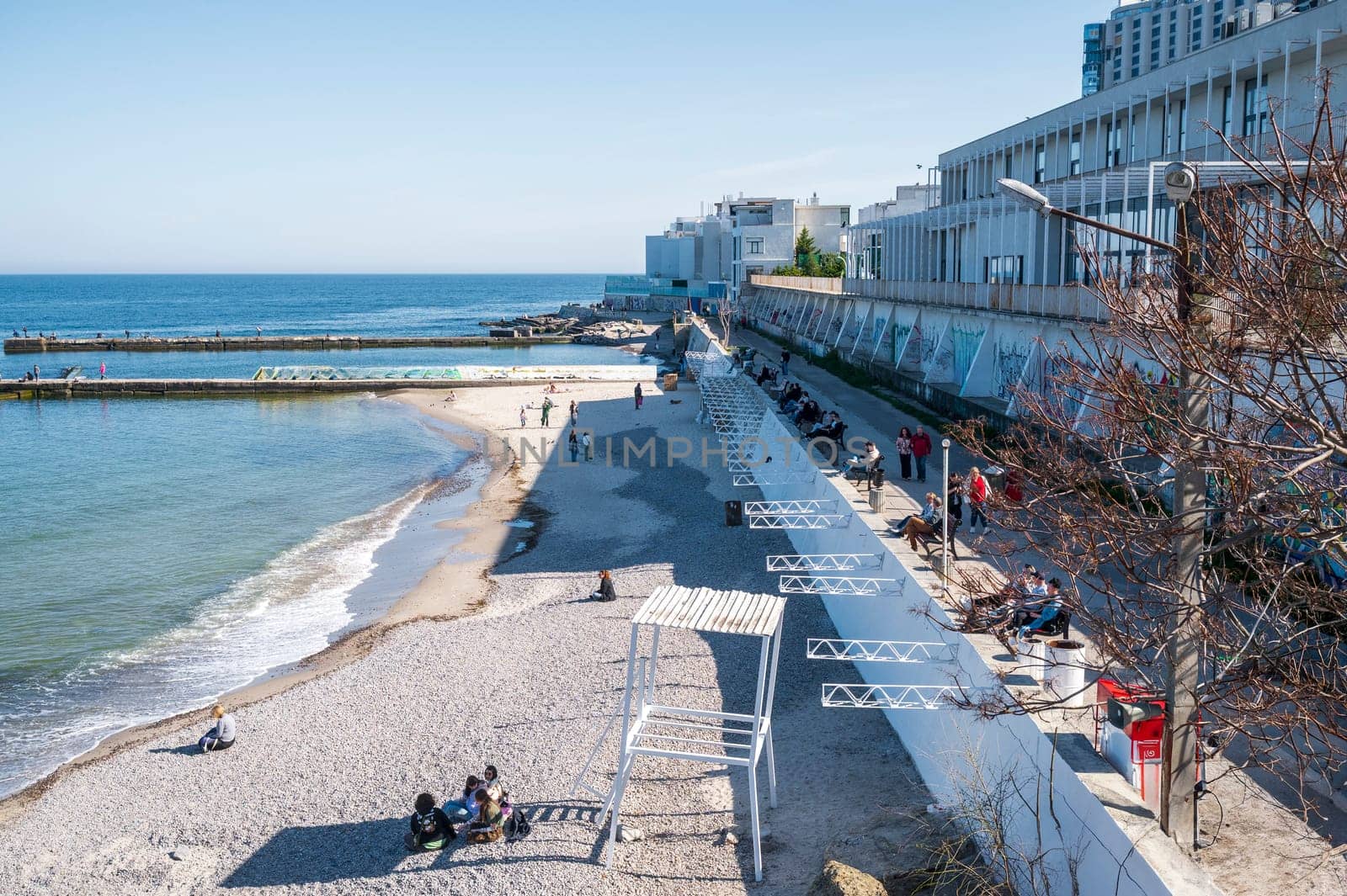 Odessa, Ukraine - April 19, 2025: City beach in Arcadia. Embankment, sea sand beach, people sitting on the beach and walking. Sandy beach in the midday sun. Concrete piers go into the calm sea.