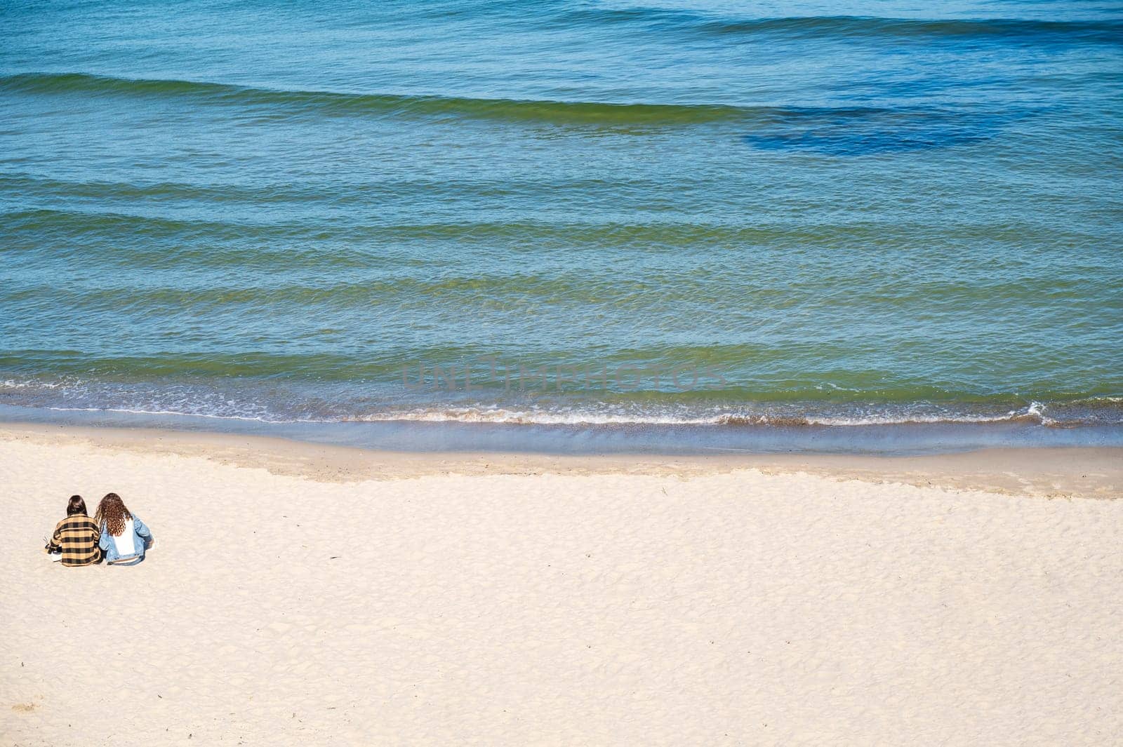 Two girls in clothes are sitting on a sandy beach and looking at the sea. Clear blue sky, calm sea. The girls are sitting with their backs to the camera. A group of people on a beach.