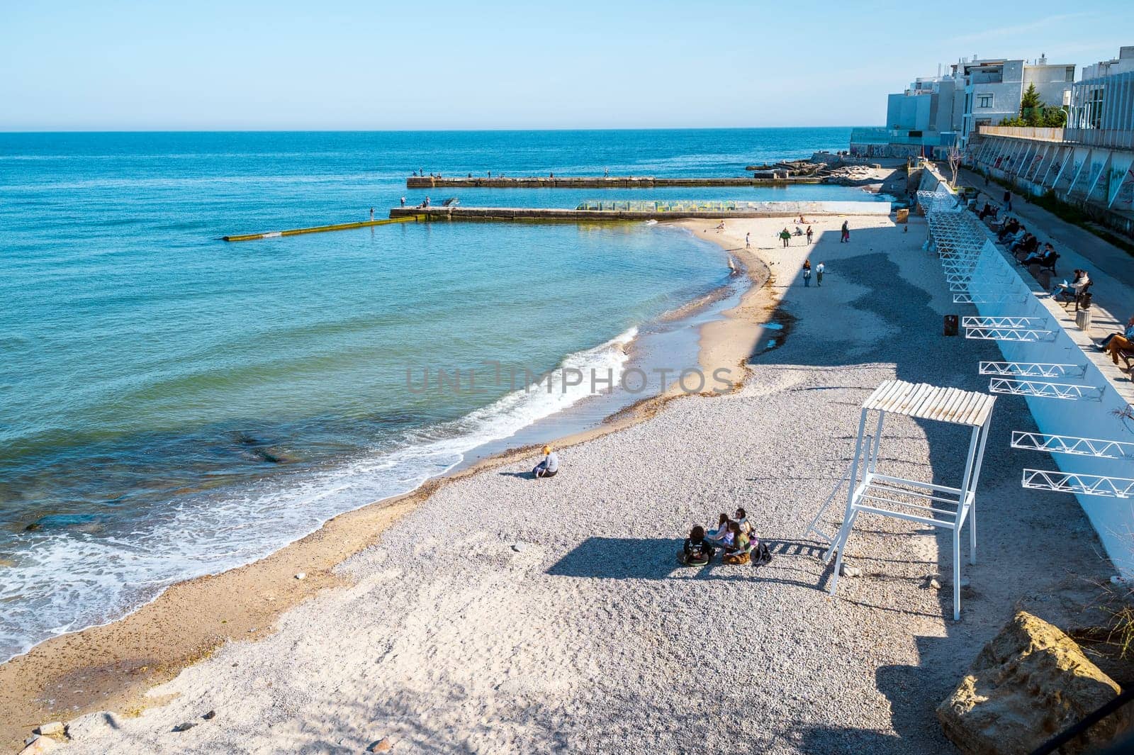 Embankment, sea sand beach in European city, people sitting on the beach and walking. Sandy beach in midday sun. Concrete jetties go into calm sea.