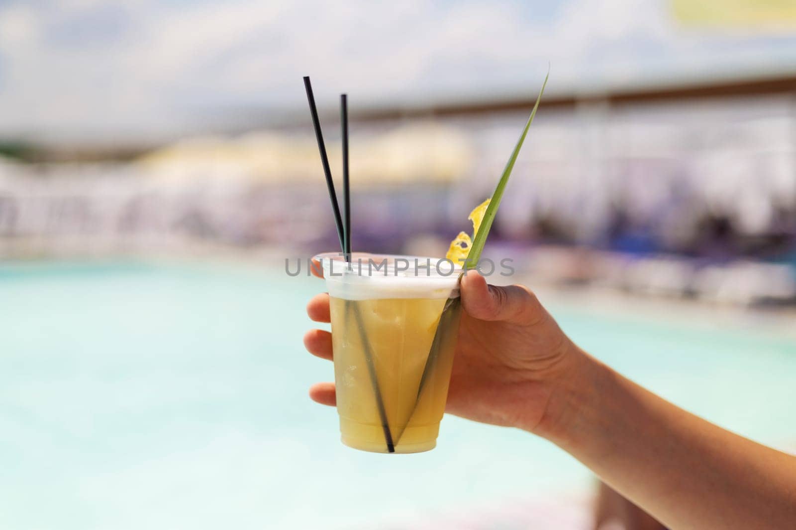 A hand holding a refreshing tropical cocktail in a plastic cup with straws and garnish, set against the backdrop of a sunny poolside. Perfect for summer vacation and leisure themes.