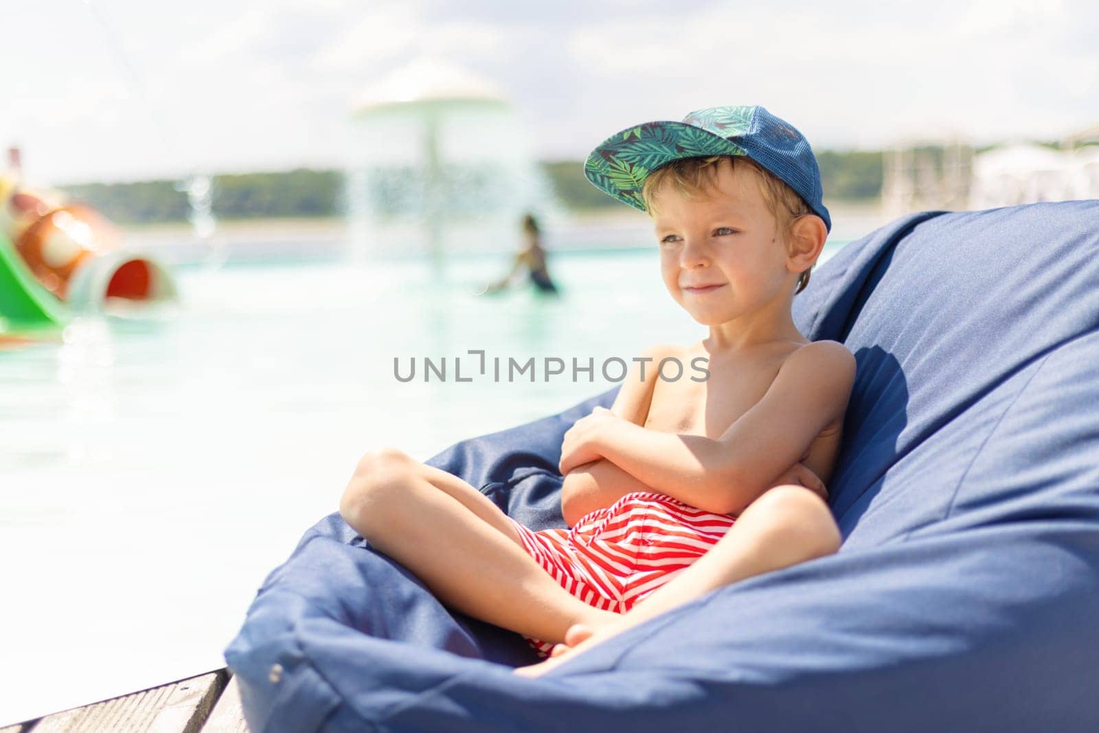 A young boy wearing a cap and striped shorts relaxes on a bean bag by the poolside on a sunny day. The background features a pool with a water fountain and other children playing, creating a cheerful summer atmosphere.