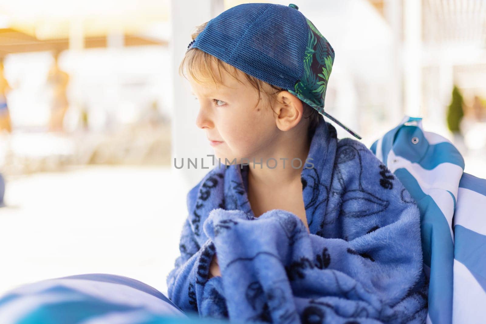 A young boy wearing a blue cap and wrapped in a cozy blue blanket, gazes thoughtfully into the distance at a sunny beach setting. The soft focus background suggests a relaxed and warm atmosphere, perfect for a summer day.