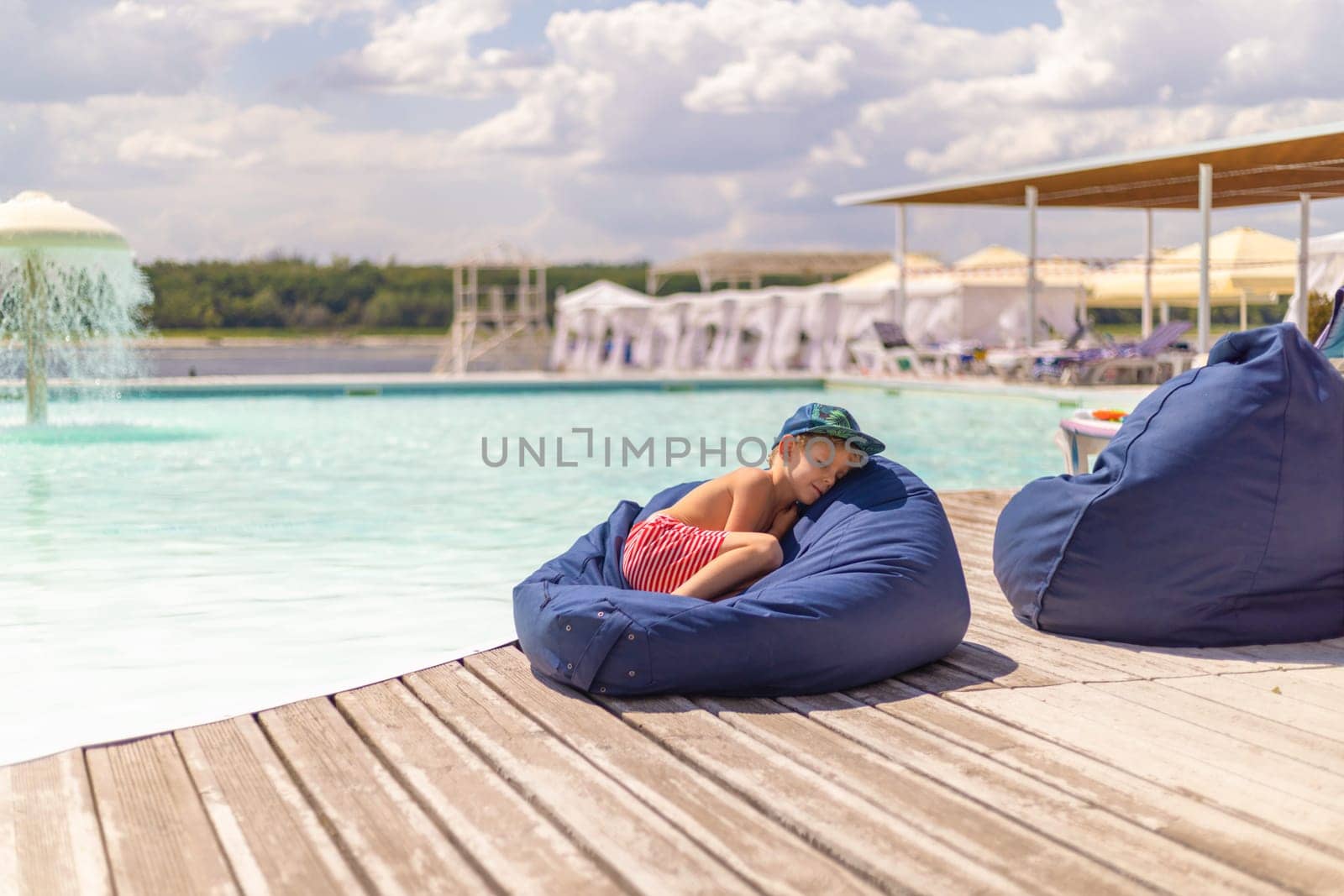 A young child wearing a cap and striped shorts is peacefully napping on a blue bean bag next to a swimming pool. The scene captures a tranquil summer day with clear skies and a serene poolside atmosphere.