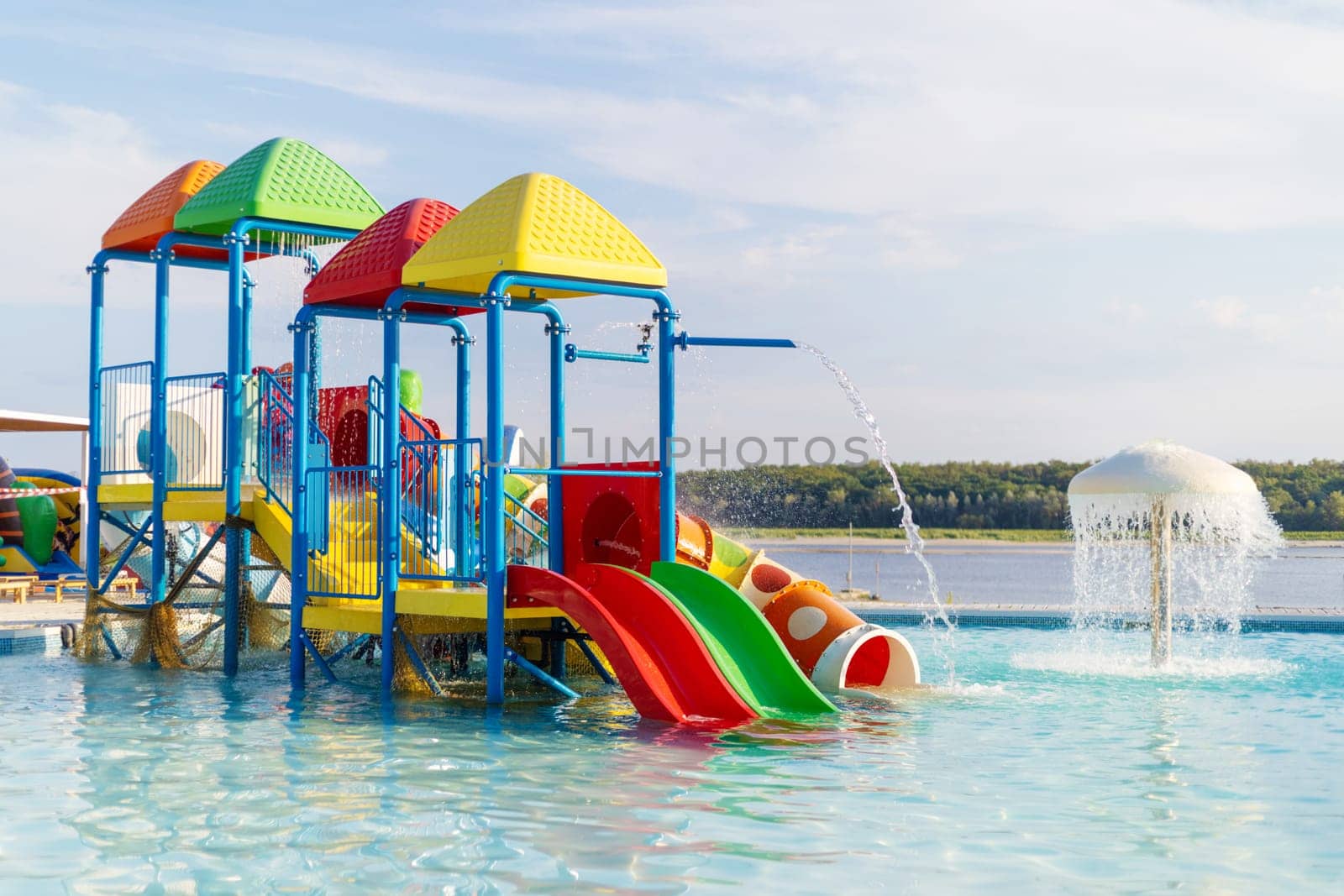 Brightly colored water playground structure with slides and water streams in a shallow pool near a lake under blue sky.