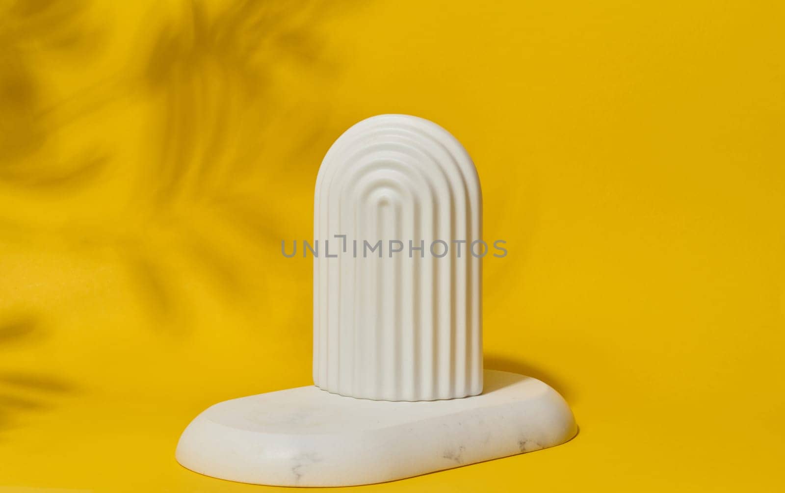 Small, white ceramic decorative arch with vertical ribbed texture stands on a bright yellow surface with blurred leaf shadows.
