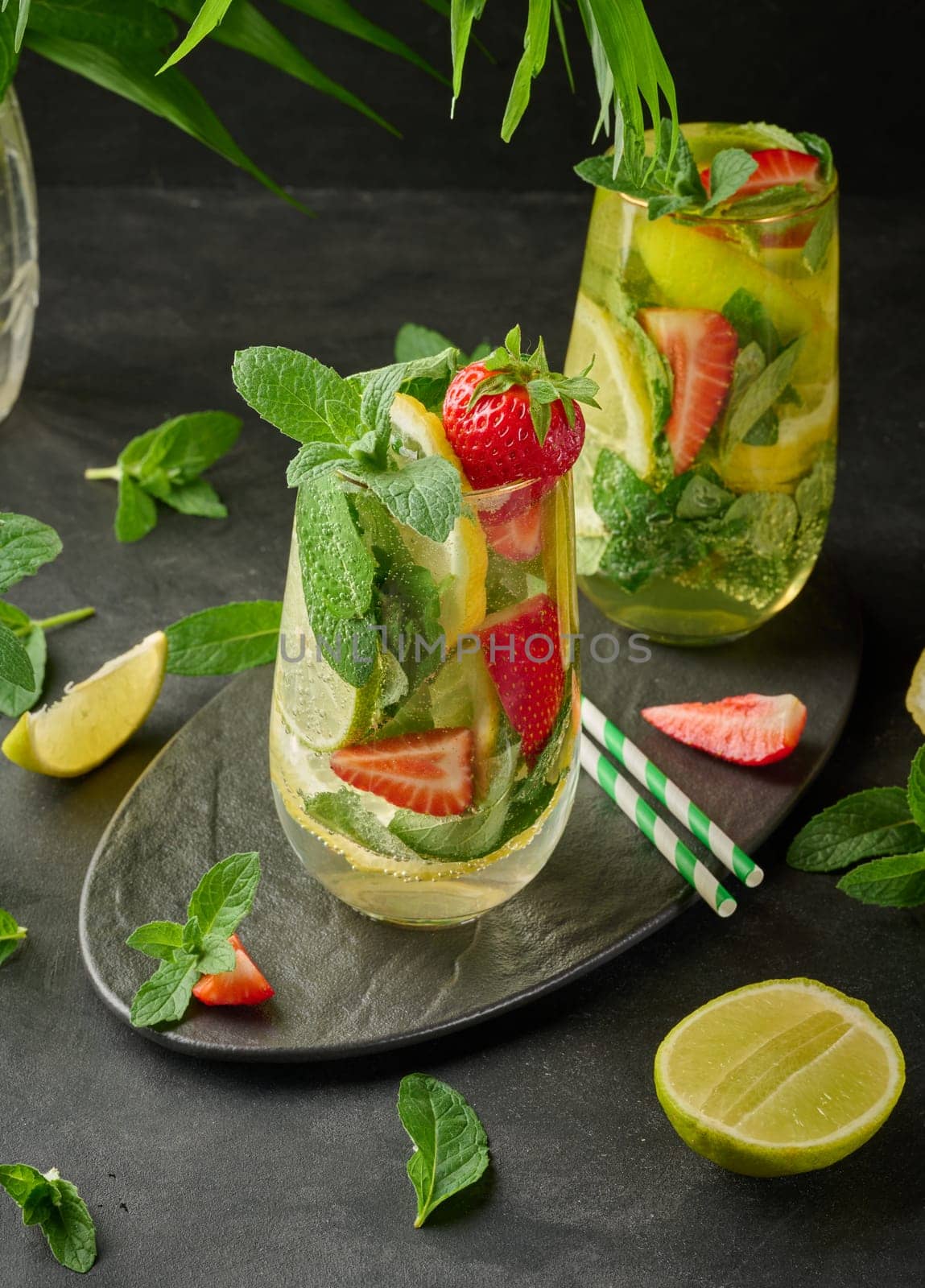 A refreshing drink in a glass with lemon slices, strawberries, fresh mint leaves, and sparkling water, served with lime and mint on a black plate