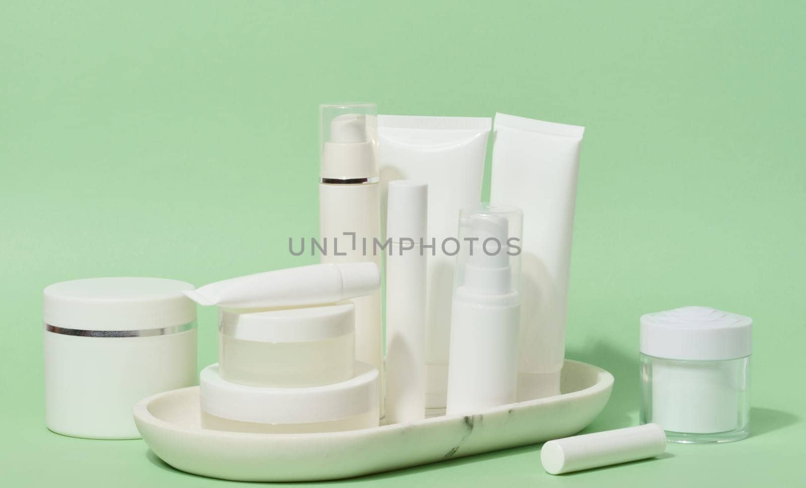A collection of various white cosmetic containers, including tubes, jars, and bottles, are arranged on a white stand against a light green background 