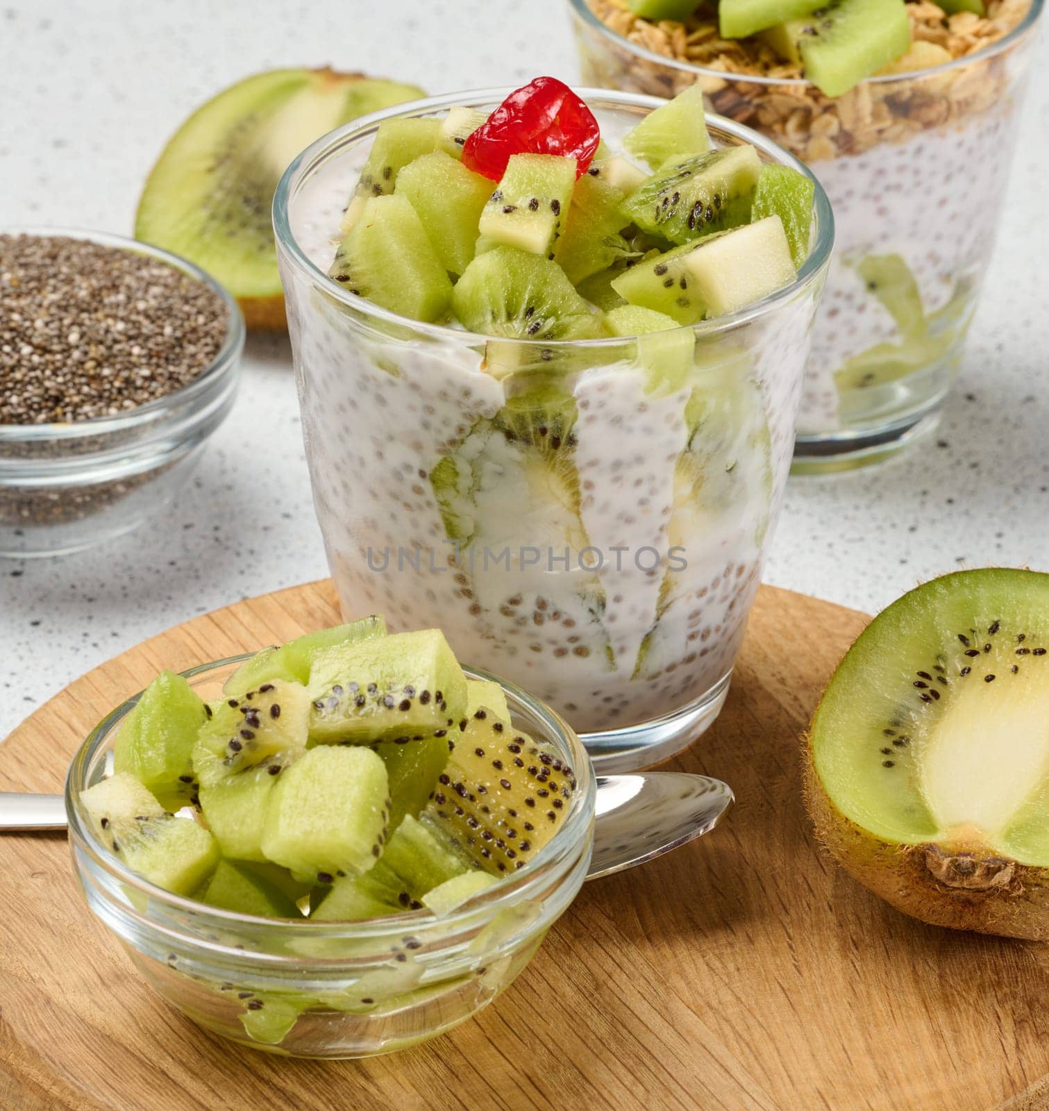 A glass of chia seed pudding layered with kiwi and granola, topped with kiwi slices, sits on a wooden board with ingredients around it