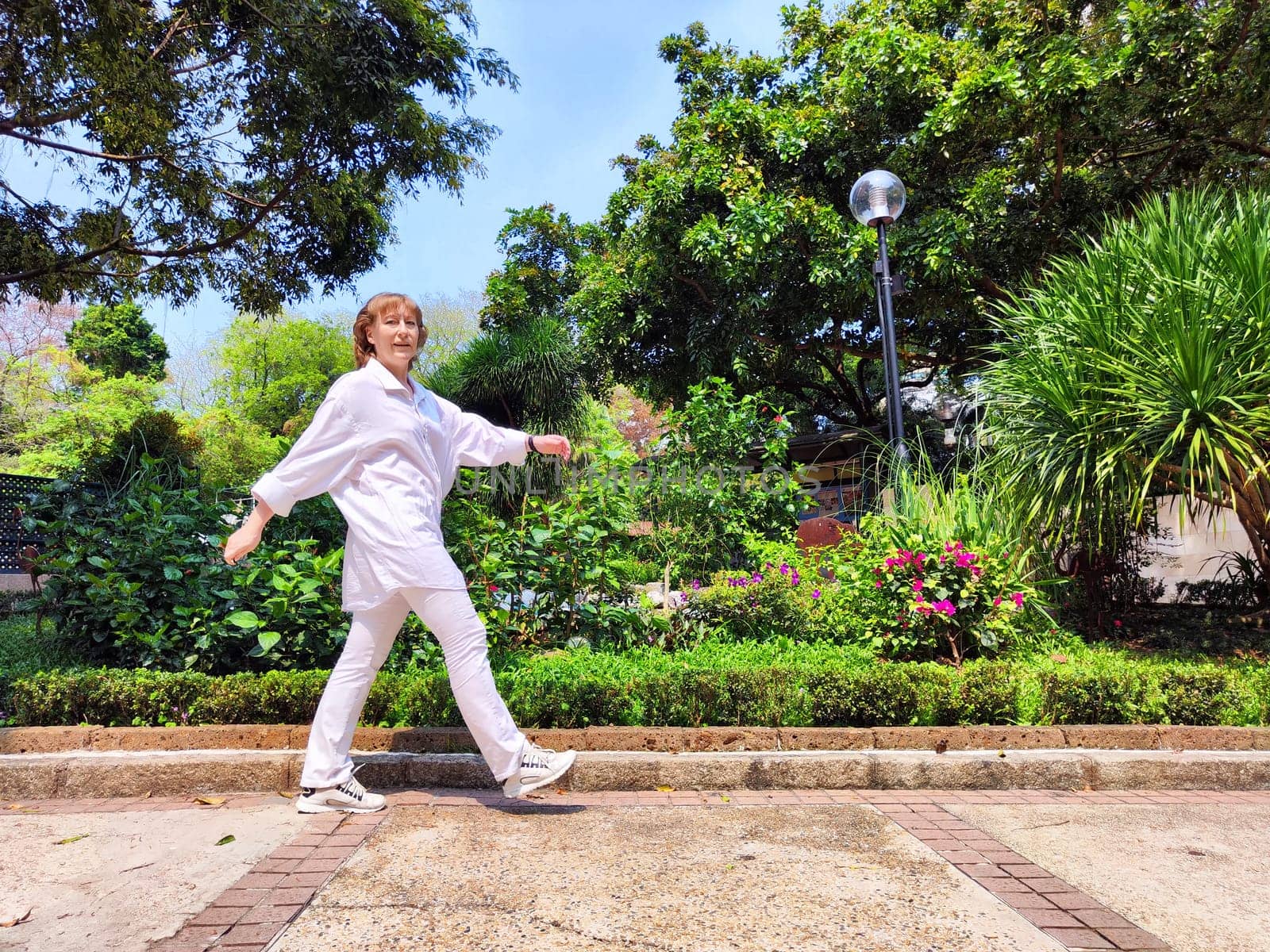 Mature woman with blonde hair walks confidently in a beautiful garden filled with greenery and colorful flowers, embracing her journey.