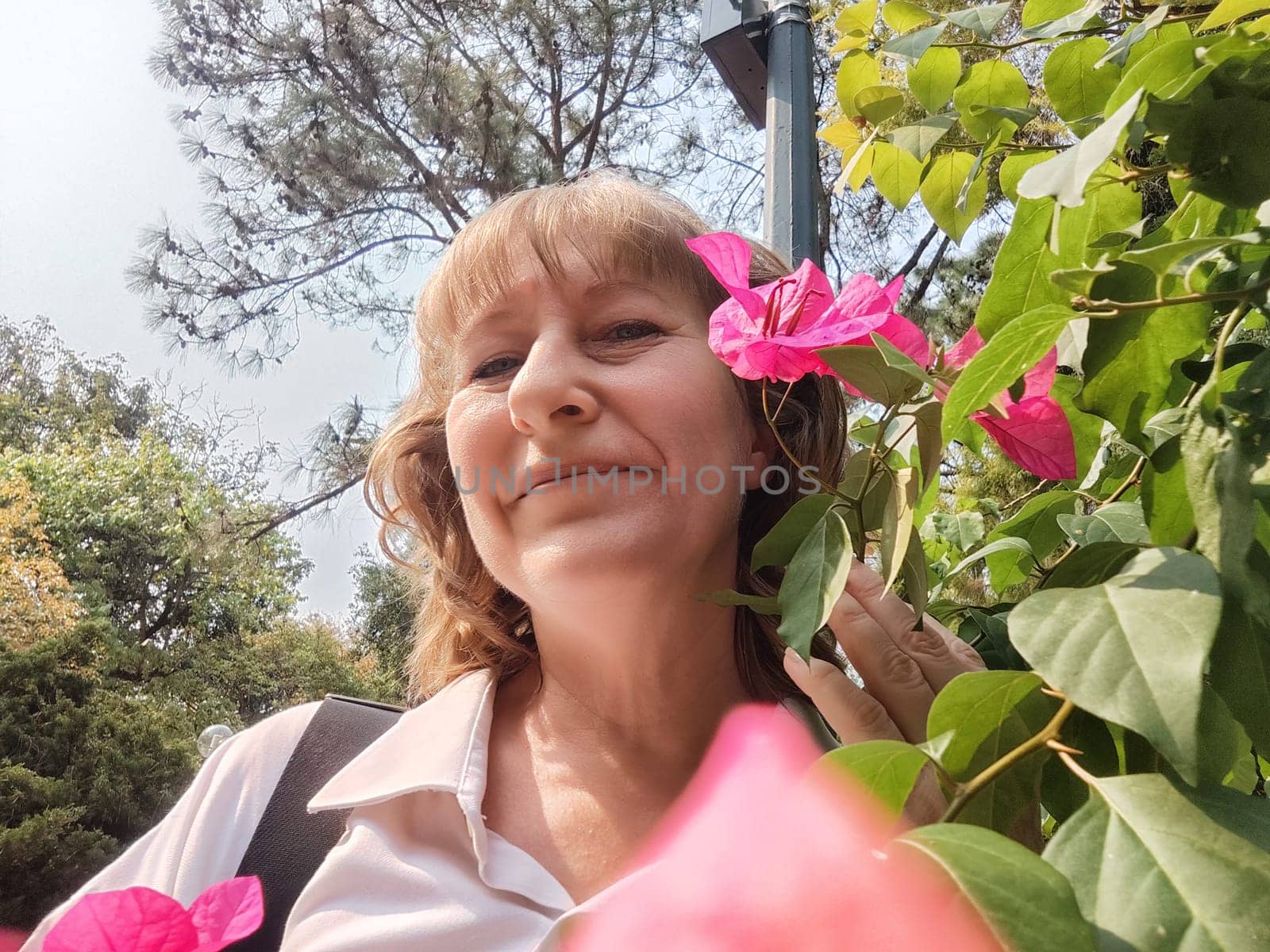 A middle-aged blonde woman smiles as she plays with vibrant pink flowers, capturing a joyful moment in a sunny garden.
