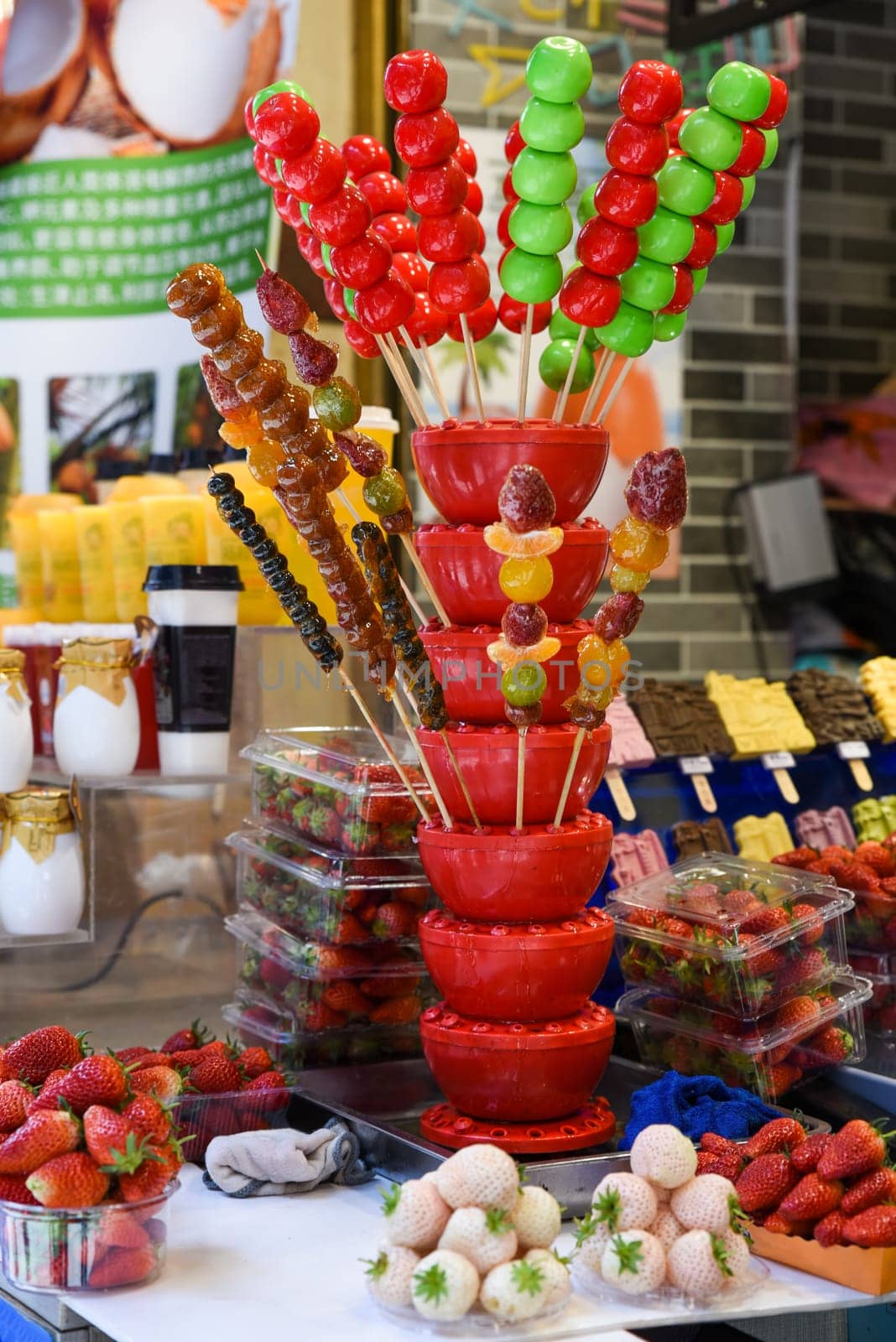 A colorful street food stall in Shanghai displays traditional Chinese candied fruits on sticks (tanghulu) - glazed apples, strawberries, and citrus arranged in vibrant pyramids alongside white strawberries and ice cream treats