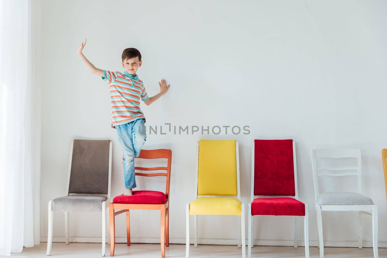 Little boy happily jumping on different chairs in white room