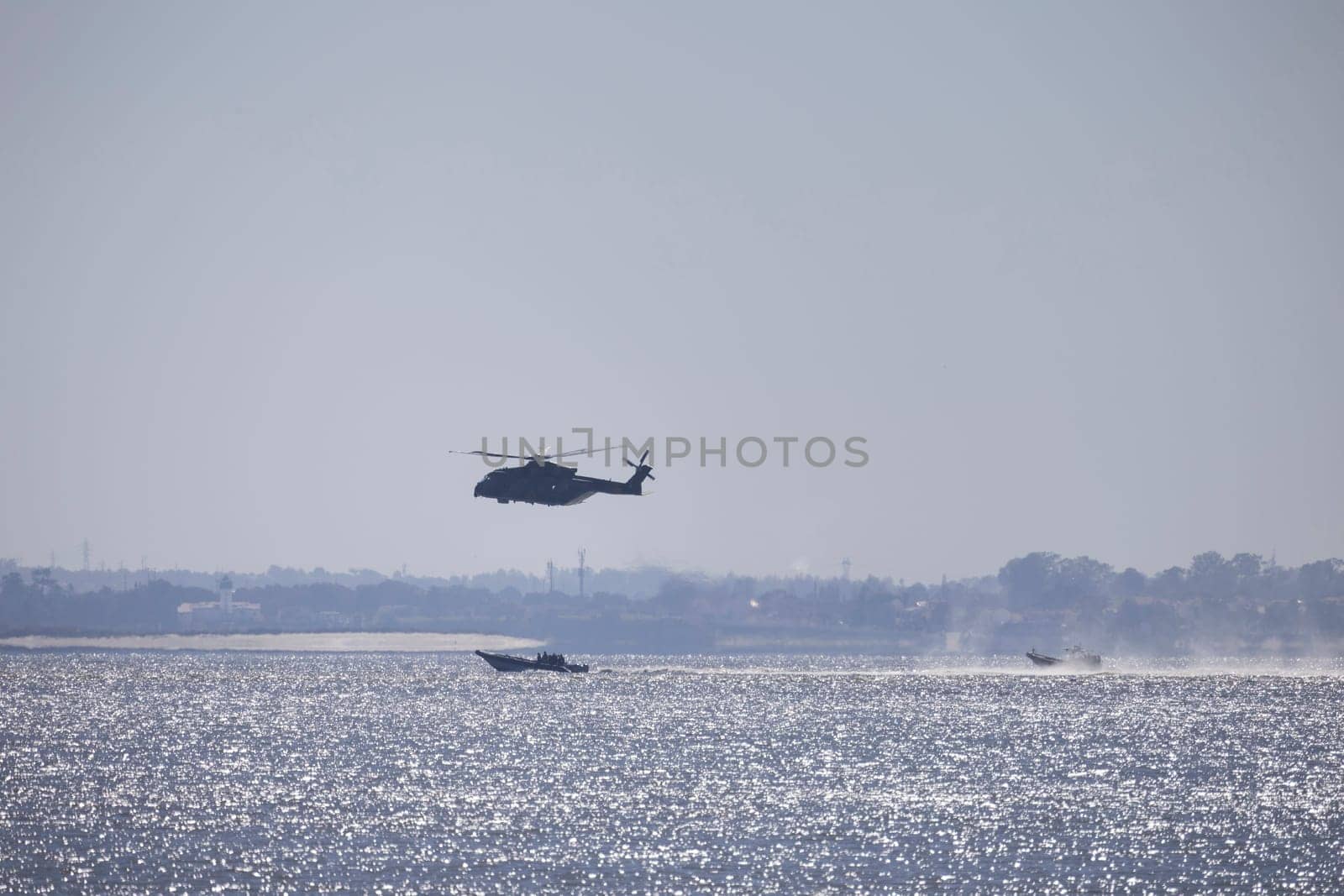Military helicopter flying over the ocean, with patrol boats assisting in surveillance and potential rescue operations