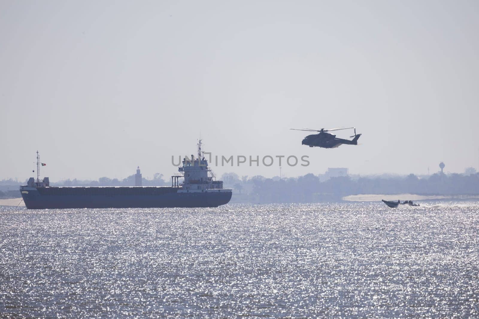 Military helicopter patrolling over cargo ship and motorboat sailing on a river