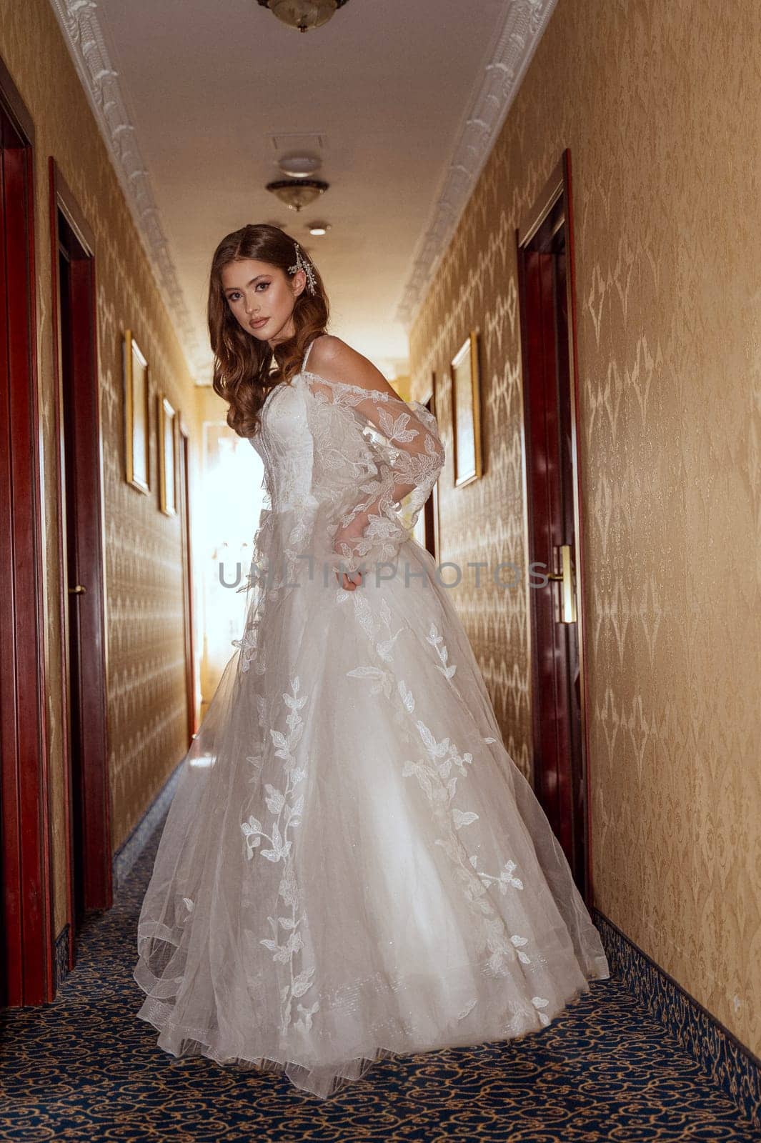 A young bride in a long classic dress with open alechi stands in the hotel corridor. She is in a silk dress with lace. She has long hair styled in a wave.