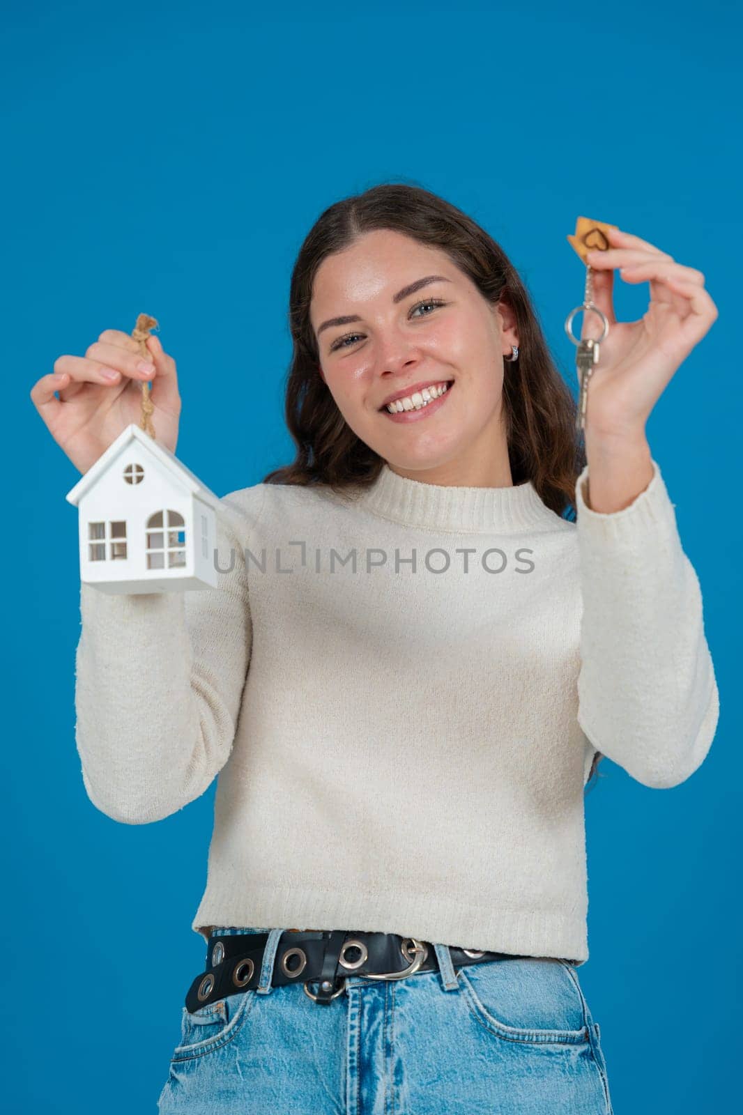 Happy young woman holding a house model and keys, expressing excitement. She wears a white sweater and stands against a blue background. Concept of home ownership, real estate, or investment.Vertical