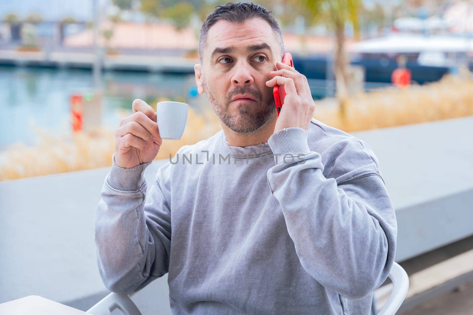 Man with short dark hair and beard drinks coffee while talking on his smartphone.He wears a casual gray sweatshirt and stands outdoors near the water, looking focused in a modern urban setting