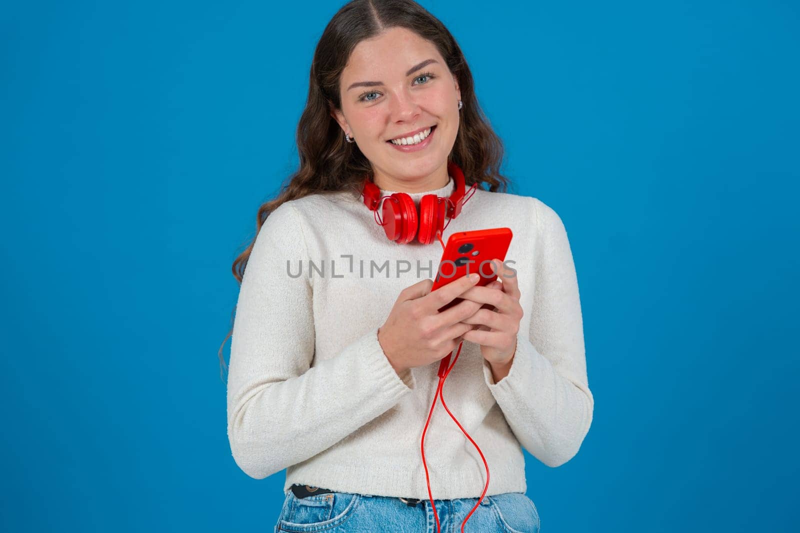 Young woman wearing red headphones around her neck, smiling while looking at her smartphone. She appears happy and excited,possibly reacting to a message or content.looking at camera, Blue background.