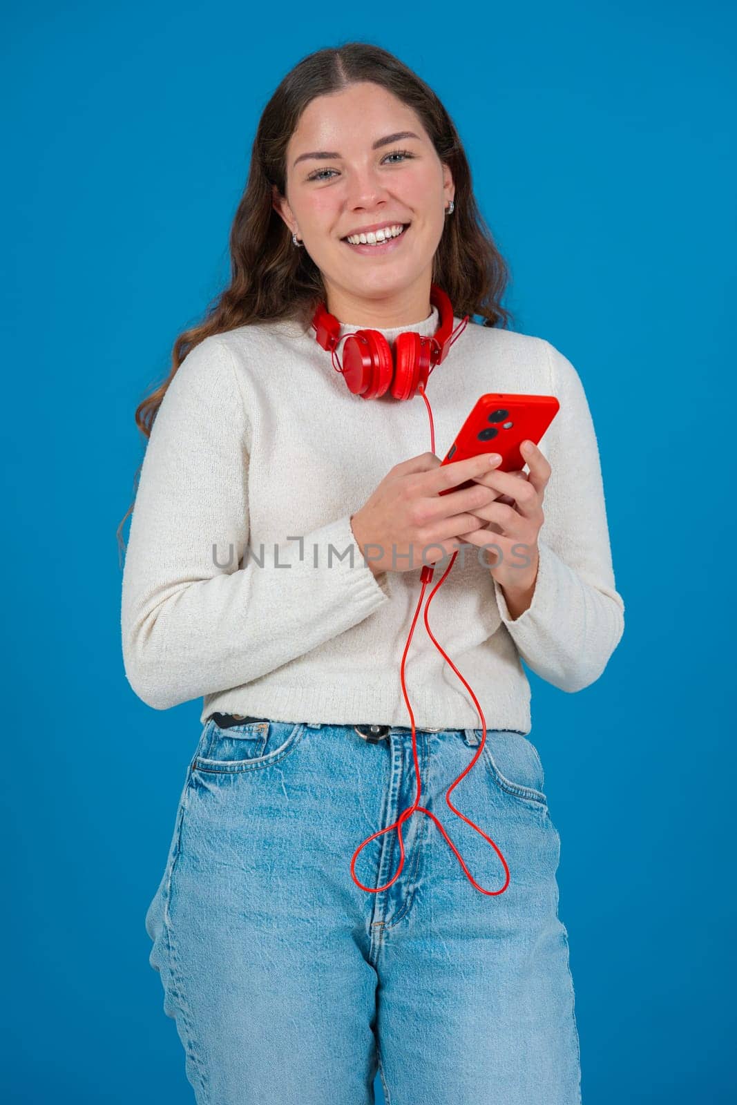 Young woman wearing red headphones around her neck, smiling while looking at her smartphone. She appears happy and excited,possibly reacting to a message or content.looking at camera, Blue background.