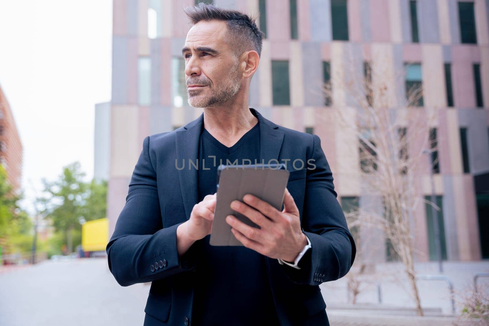 Businessman using a tablet outdoors in a modern city while looking away. He wears a black suit and smartwatch. Concept of technology, focus, mobility, and digital professional lifestyle.