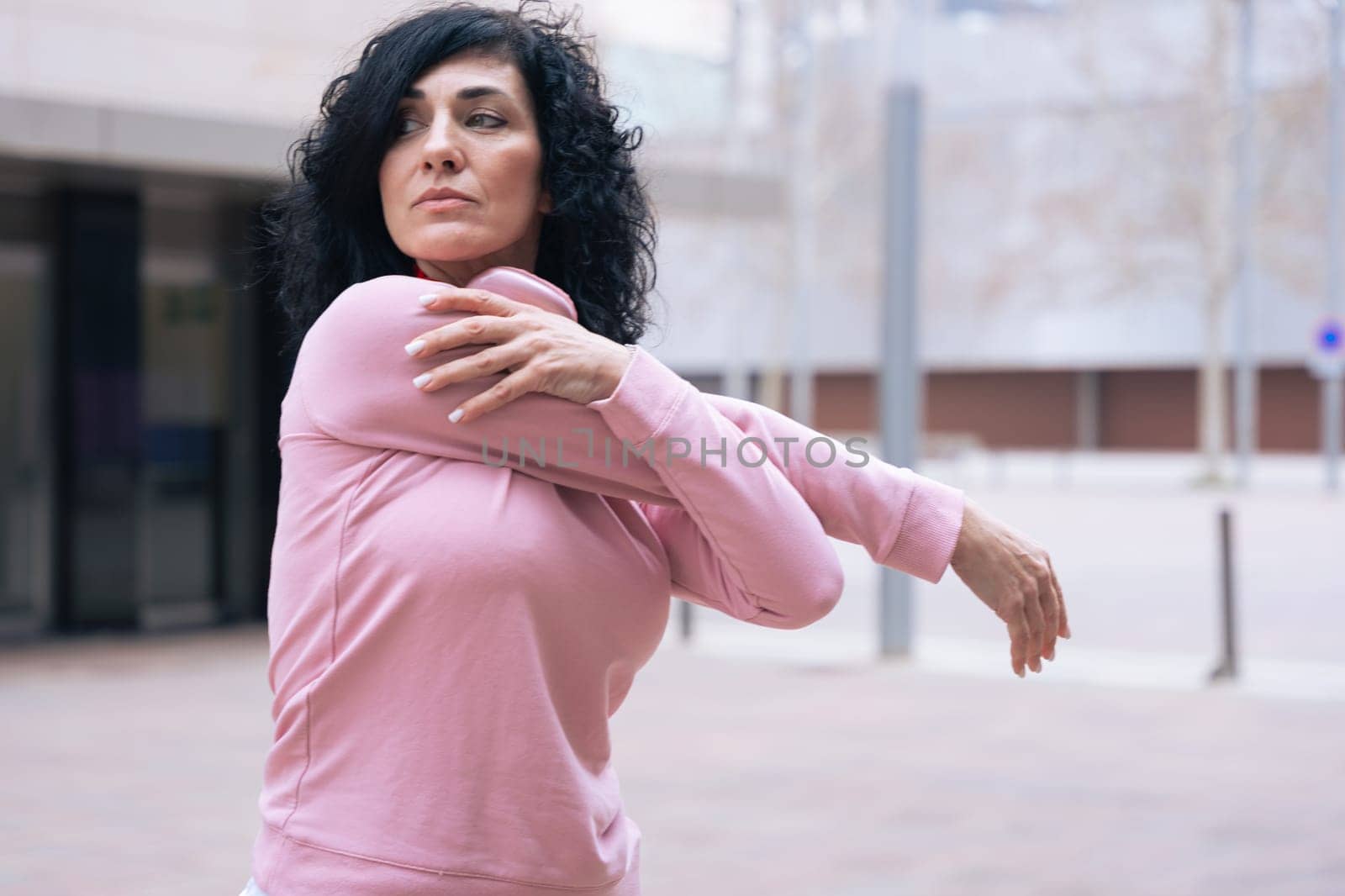 Caucasian adult woman with curly black hair stretching her arms outdoors in an urban setting.she is wearing a pink sweatshirt and exercising for a healthy lifestyle, fitness, and wellness in the city