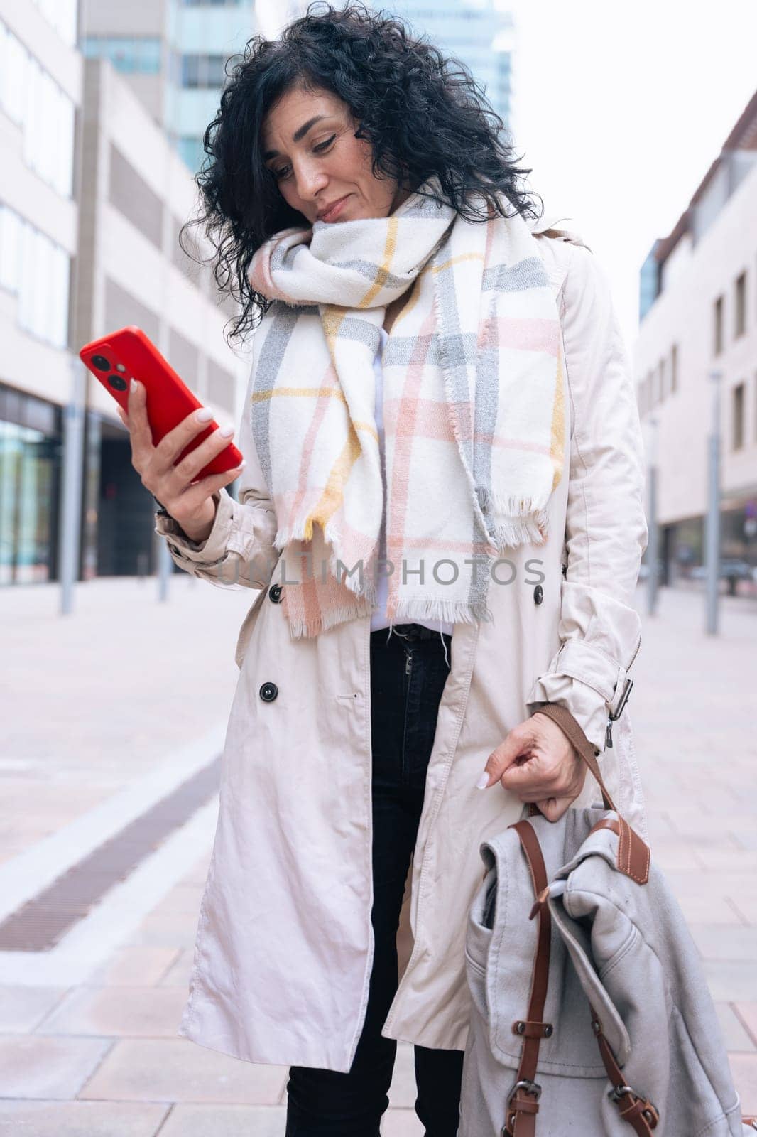 Serious caucasian woman standing outdoors in urban setting, wearing coat and scarf, using her red smartphone. She is serious and relaxed, enjoying a casual moment on a cold day in the city. Vertical