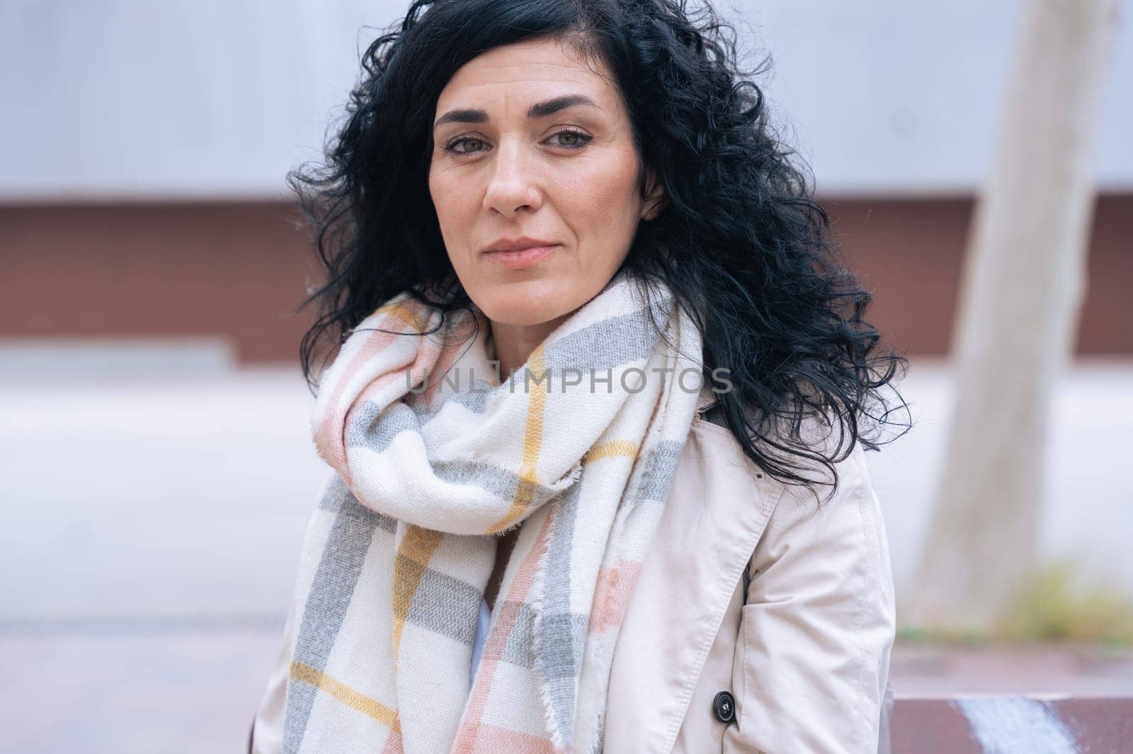 Horizontal portrait of a woman sitting on a bench outdoors. She wears a beige coat and scarf, looking calm and confident. Urban background with winter atmosphere and seasonal fashion concept.