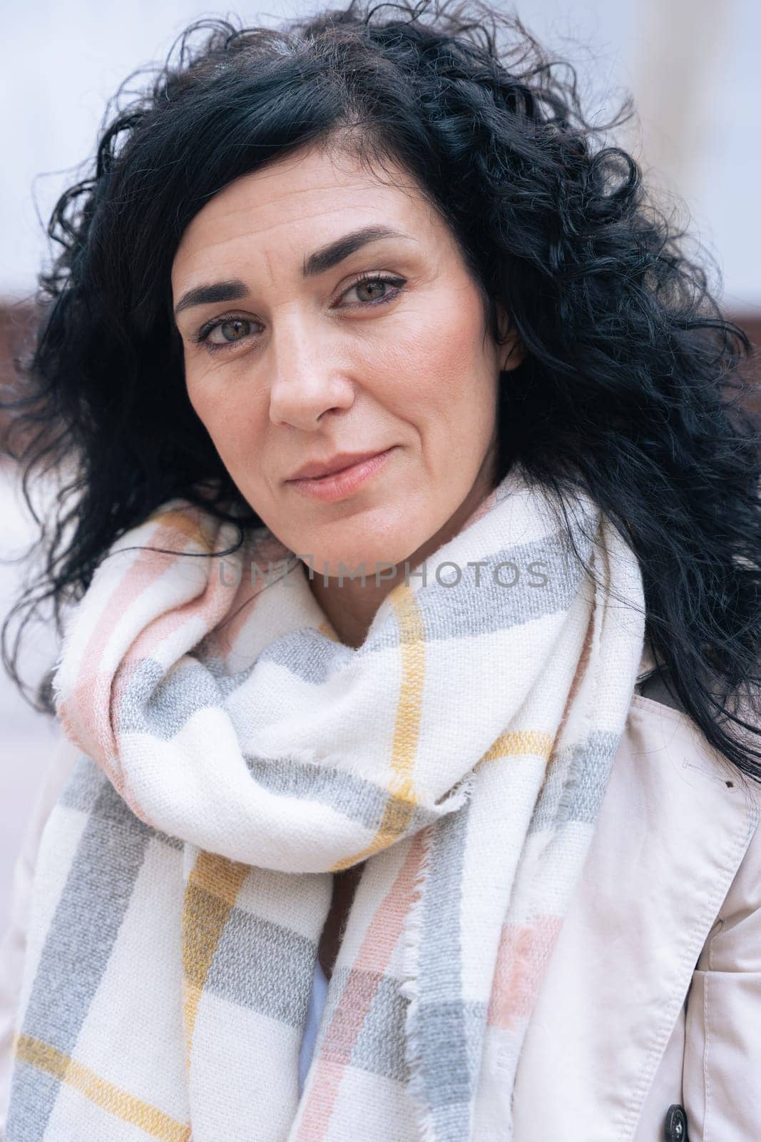 Vertical portrait of a woman sitting on a bench outdoors. She wears a beige coat and scarf, looking calm and confident. Urban background with winter atmosphere and seasonal fashion concept.