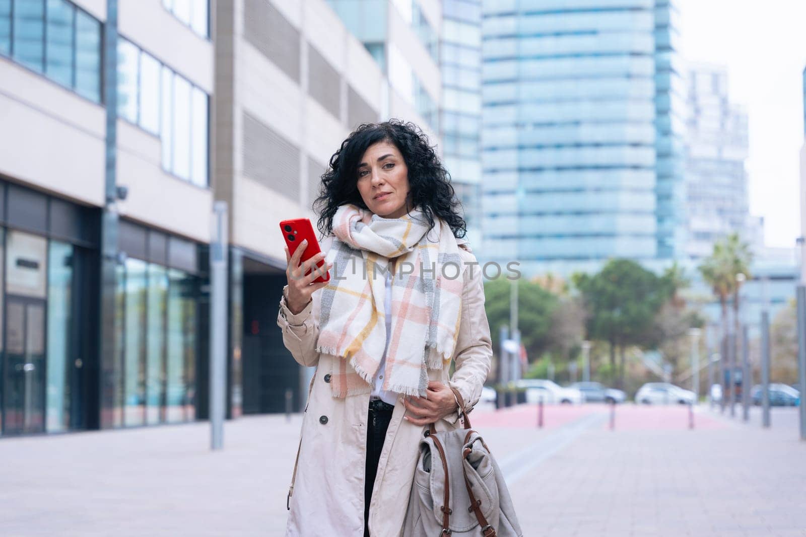 Happy caucasian woman standing outdoors in urban setting,wearing coat and scarf,using her red smartphone.She is smiling and relaxed,enjoying a casual moment on a cold day in the city looking at camera