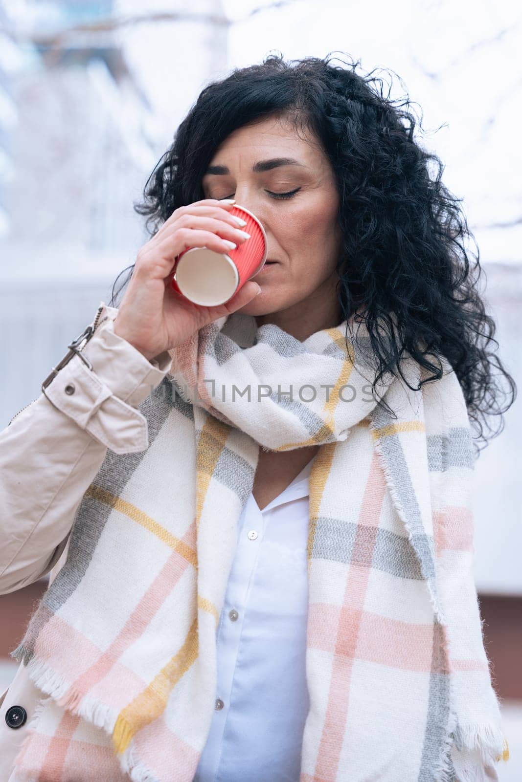 Caucasian woman enjoys a warm drink from a red cup while standing outdoors. She wears a soft scarf and light jacket, suggesting a cool day. Calm and cozy moment captured in natural daylight. Vertical