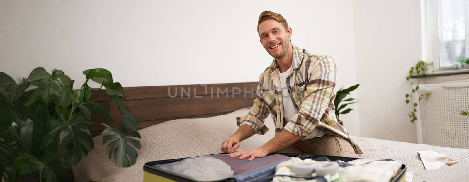 Portrait of young handsome bearded guy, tourist going on holiday, fastening his clothes in suitcase, packing for vacation, going on business trip, sitting in bedroom.
