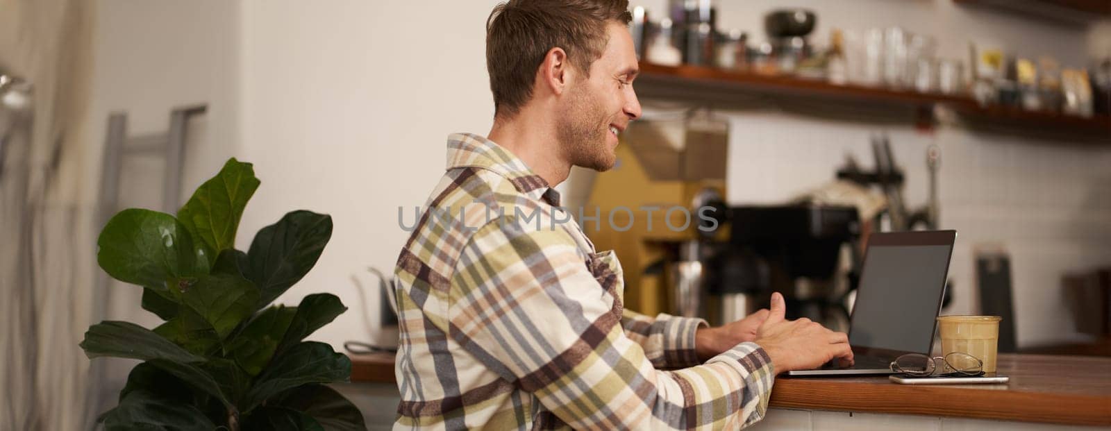Portrait of handsome smiling man, sitting with laptop in a cafe, typing, working remotely from co-working space, drinking coffee, freelancing from public space.