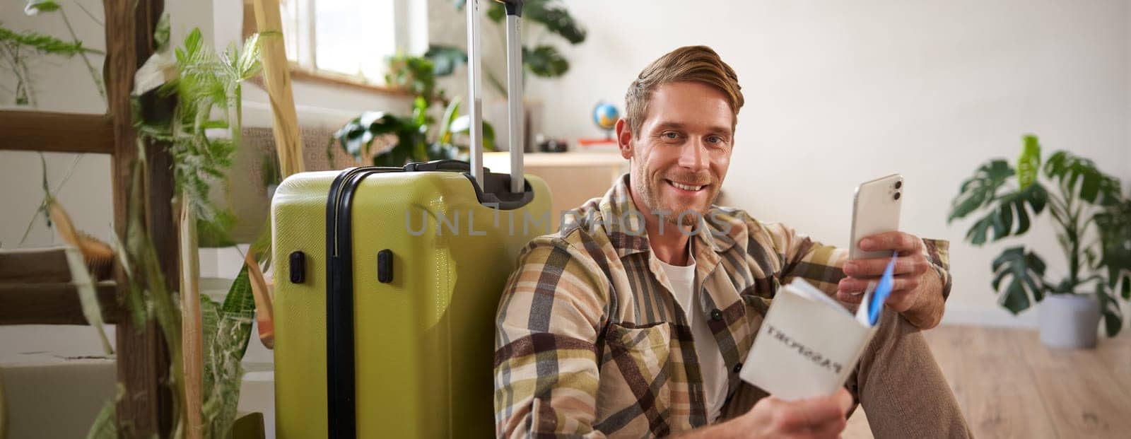 Portrait of smiling tourist, man sits with suitcase, holds smartphone, passport and tickets, goes on holiday. Tourism and people concept