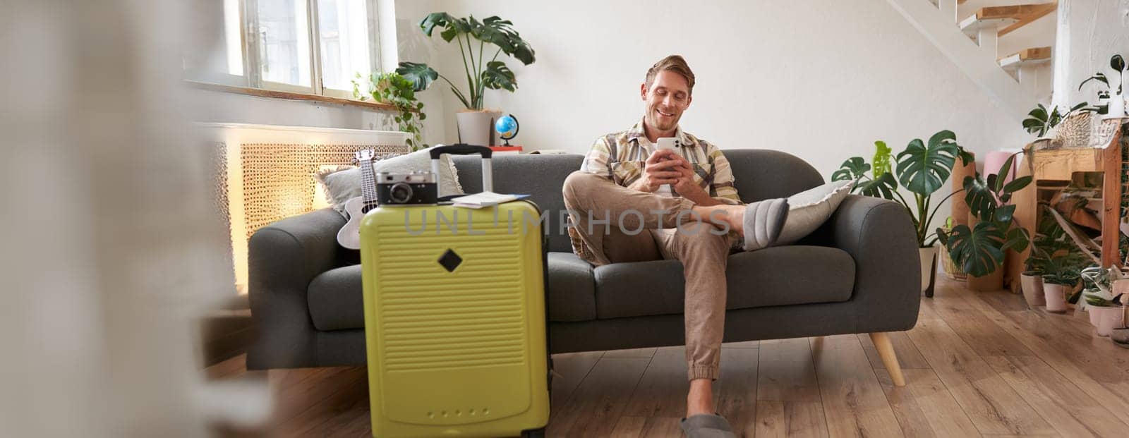Handsome young man sits on sofa with suitcase, holds smartphone, books hostel or holiday vacation, chats on mobile phone. Tourism and travelling concept