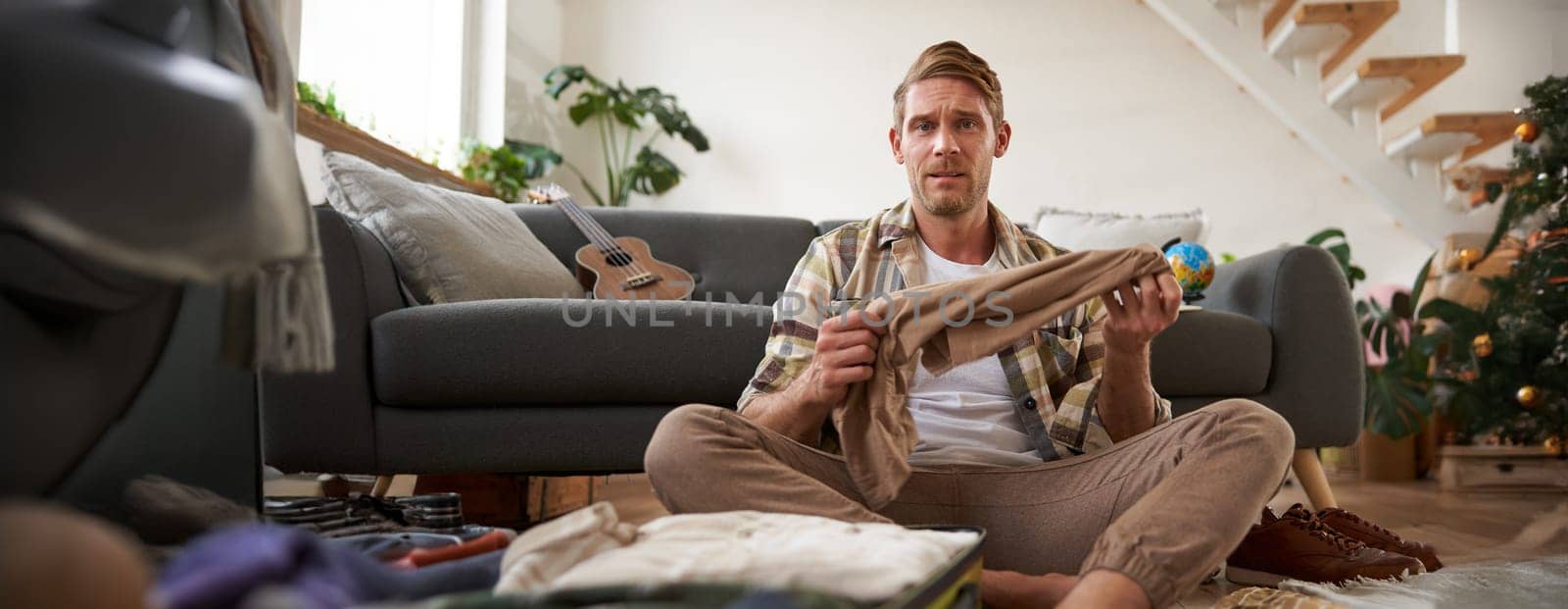 Portrait of confused man, sitting in living room with suitcase, holding pair of trousers, looking clueless, struggling to pack for holiday. Tourism and travelling concept