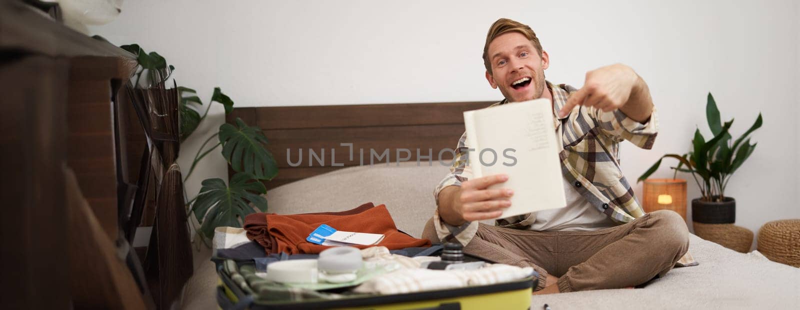 Portrait of young man pointing at his list of items, packing clothes on vacation, showing notebook, sitting near opened suitcase. Lifestyle and people concept